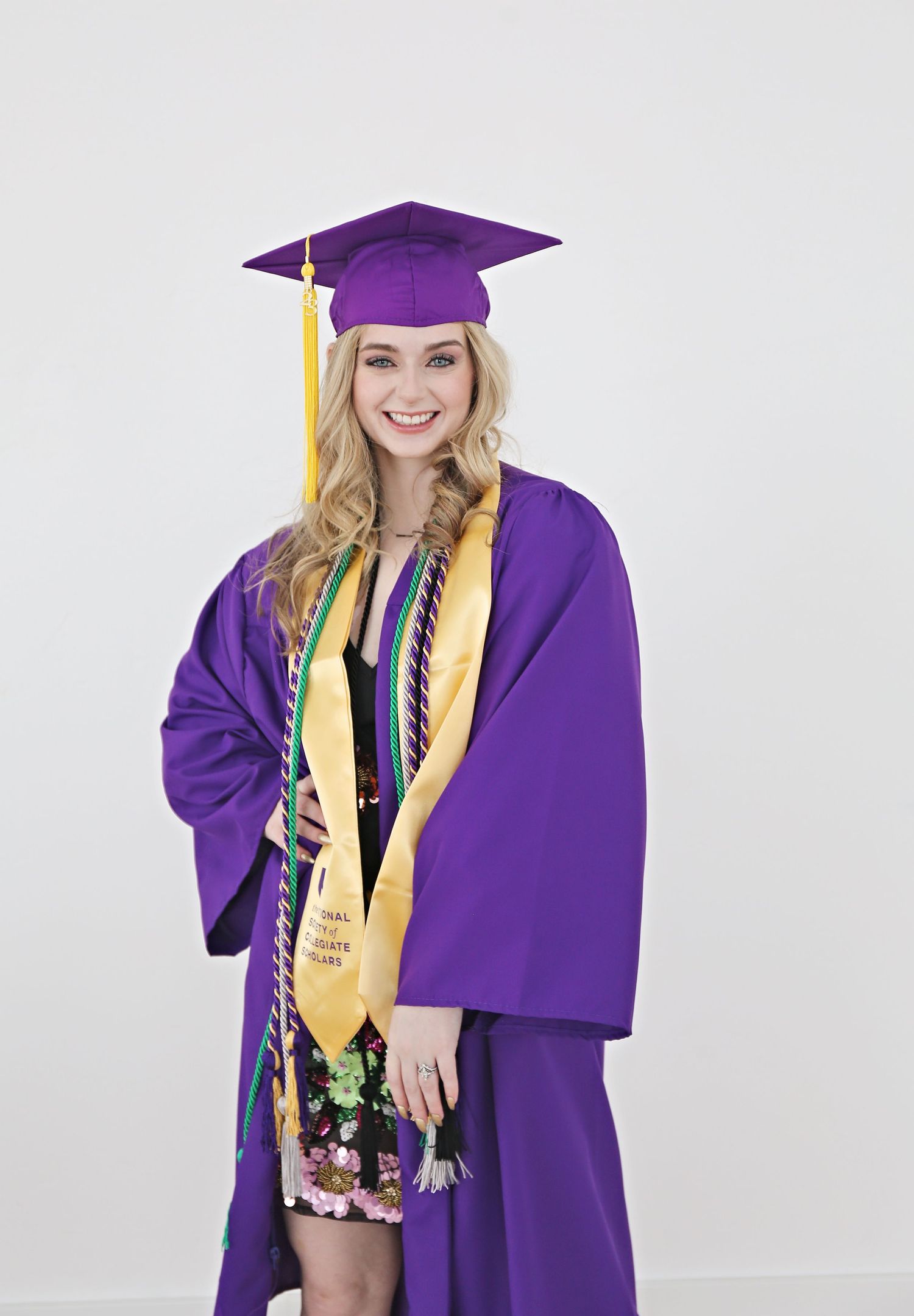 Graduate poses in purple cap and gown with gold honors cords against white background.