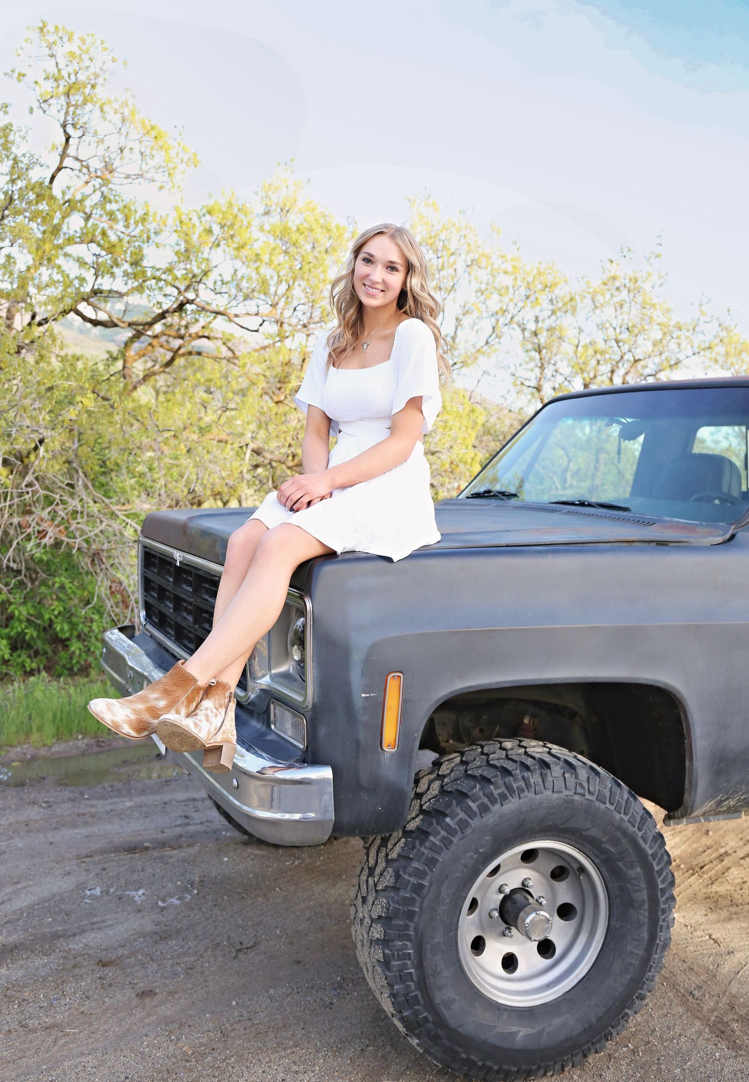 A white dress and heels outfit contrasts with a rugged lifted black pickup truck on a dirt road near blooming trees.