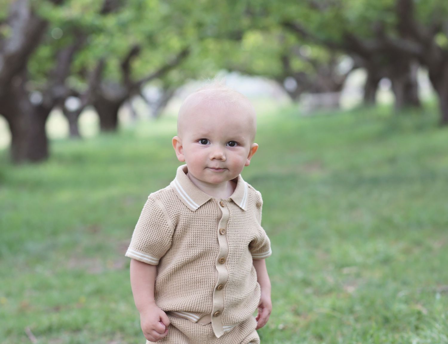 Small child in tan button-up shirt and dark pants stands in a grassy outdoor setting.