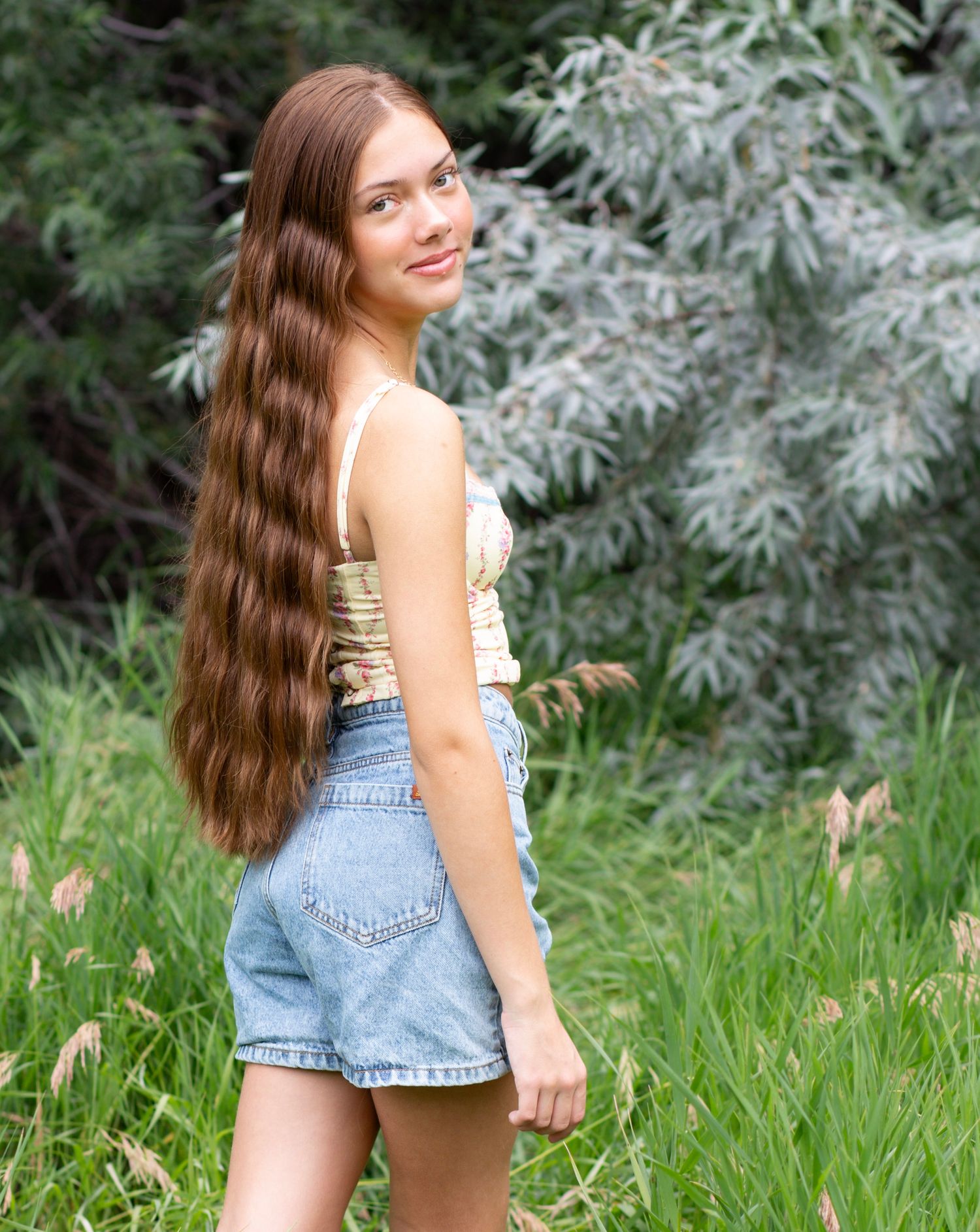 Long-haired model in denim shorts and crop top poses in front of lush green foliage.