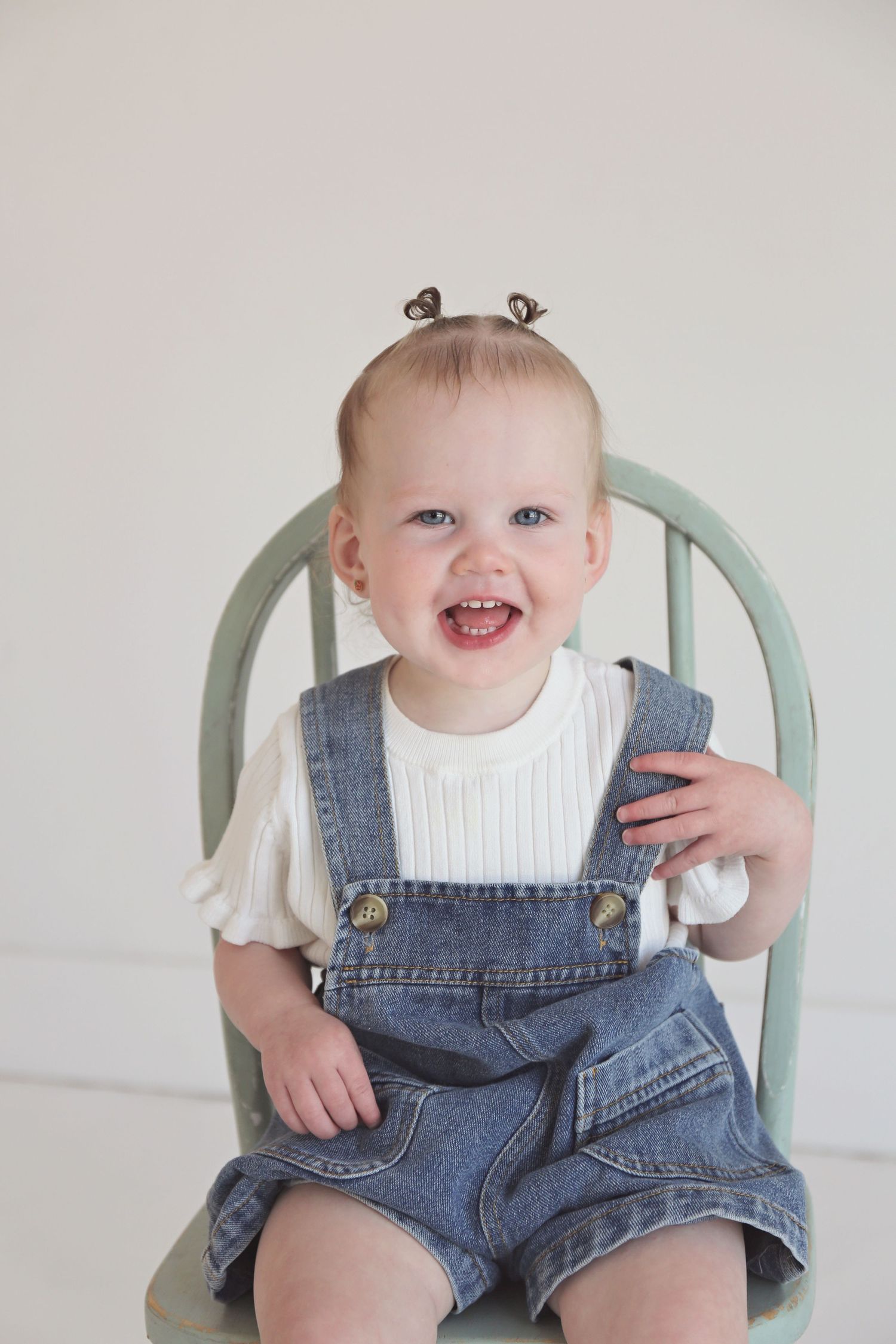 Happy baby wearing jean overalls and white shirt sits in painted wooden chair.