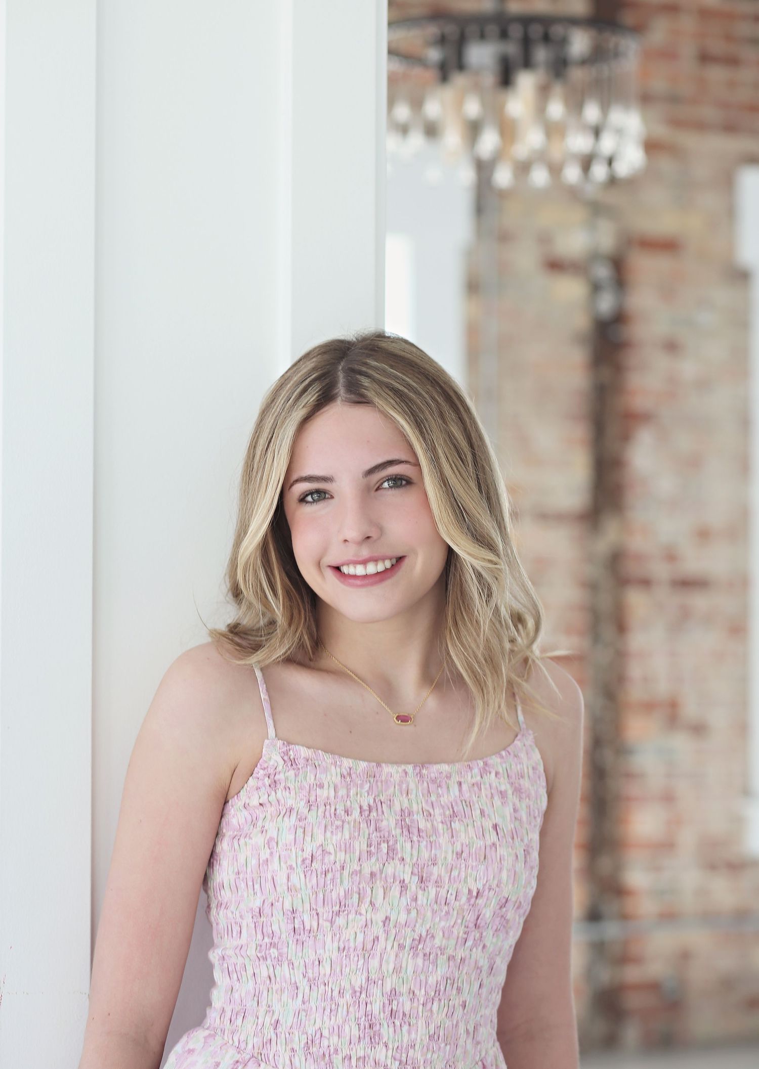 A portrait in a pink floral dress against a white wall with exposed brick and chandelier.