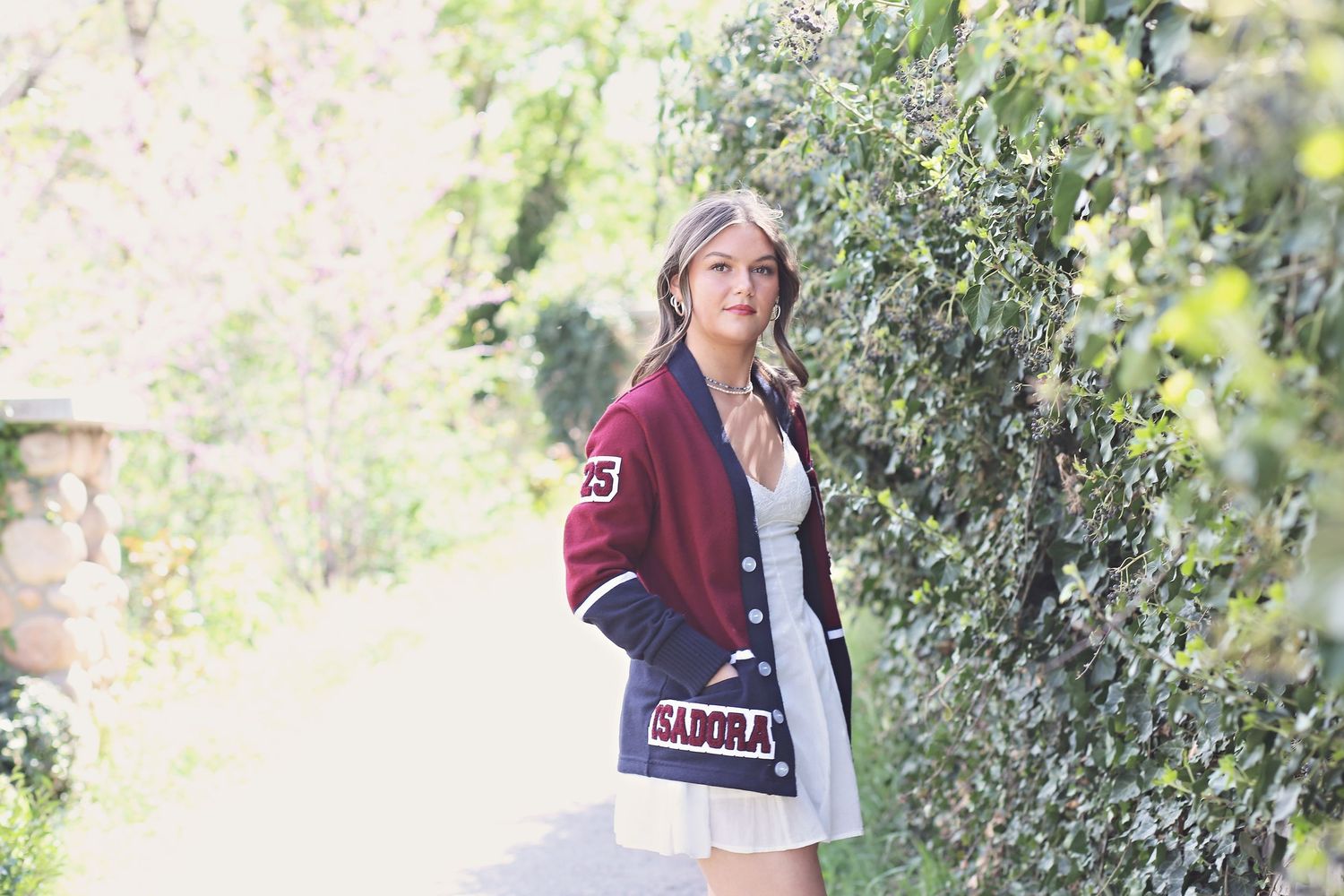 Person wearing varsity jacket and white dress on nature path surrounded by greenery.