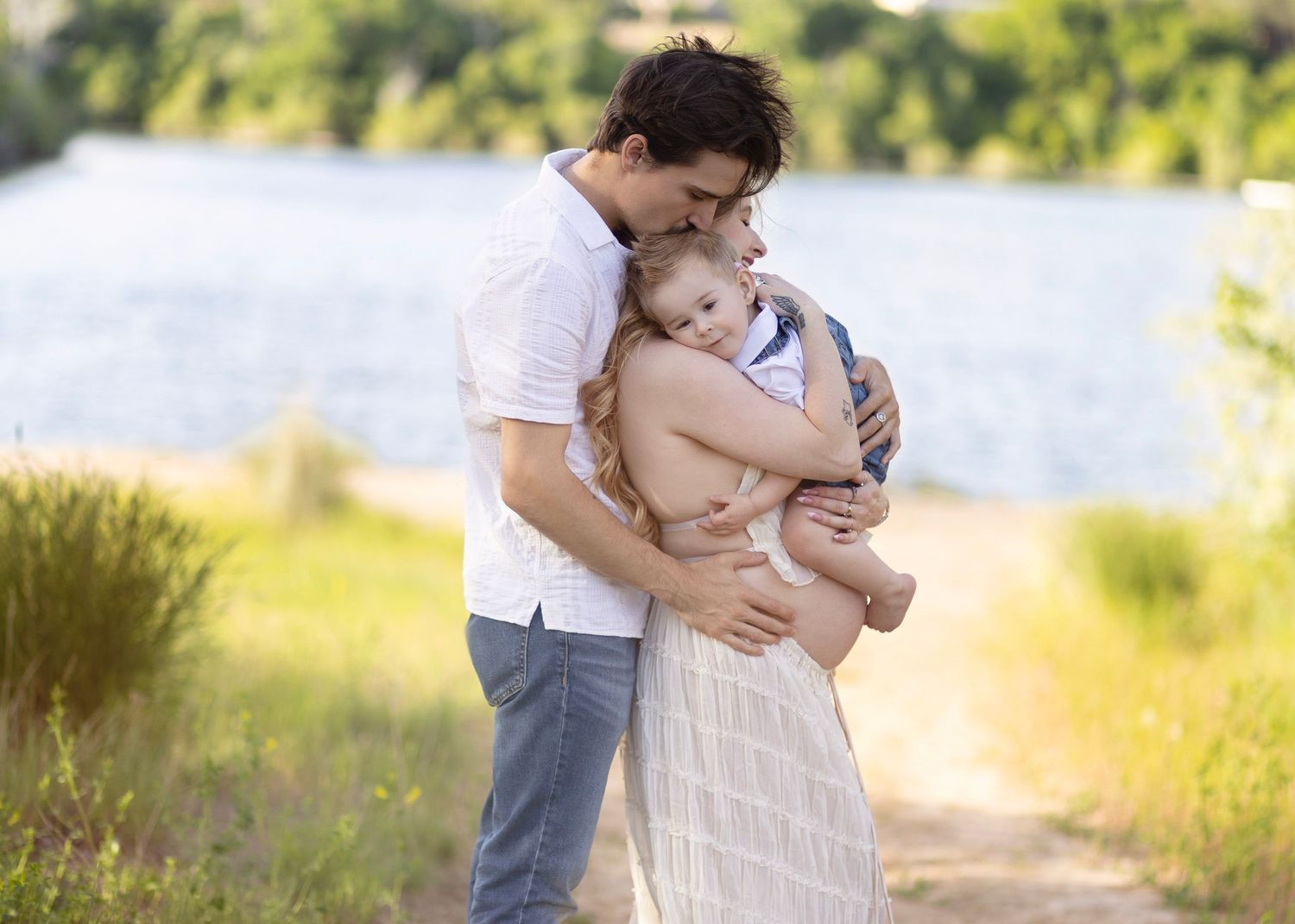 A father tenderly embraces his young child outdoors near a lake, surrounded by lush greenery on a sunny day.