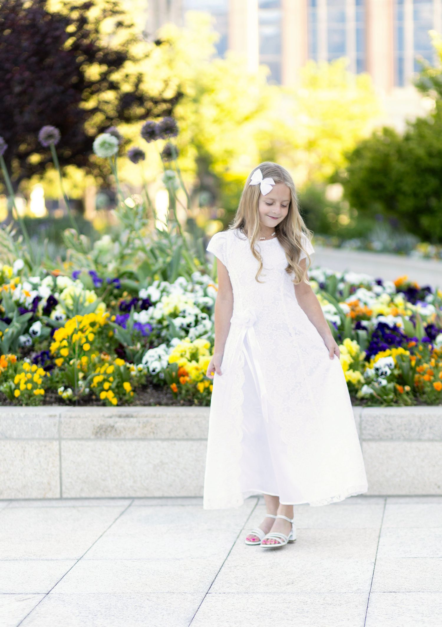 Young girl in white dress standing near colorful flower garden on sunny day.