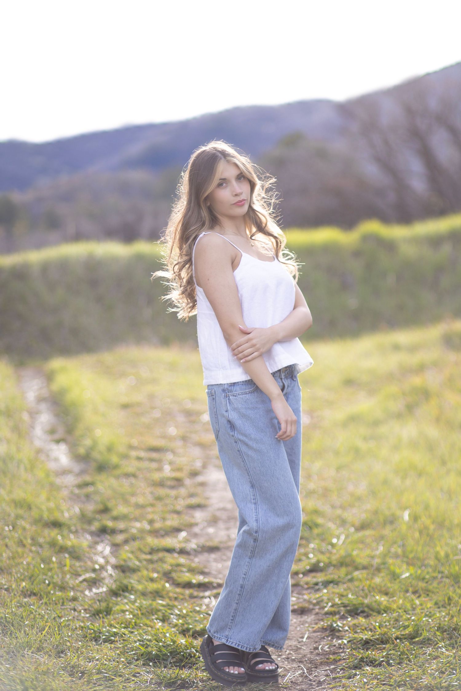 Young woman in white cami and wide-leg jeans standing on a rural path with mountains in the background.