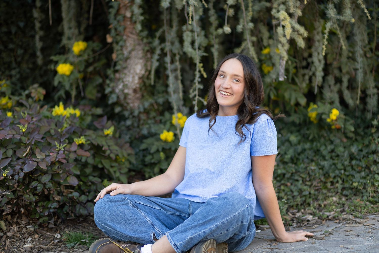 Young woman in blue t-shirt and jeans sitting cross-legged outdoors, smiling, with green foliage background.