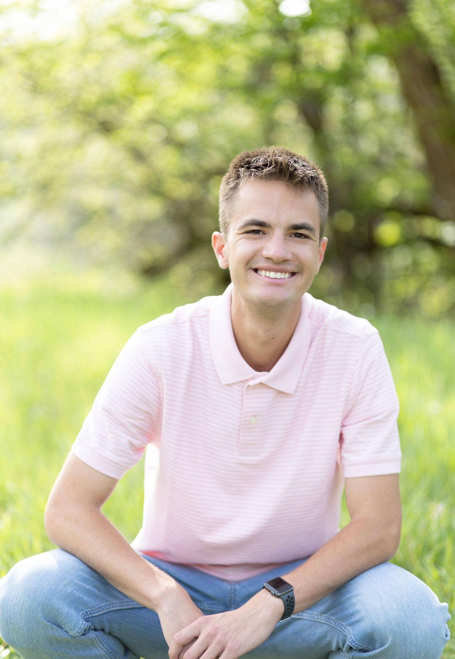 Young man in pink polo shirt sitting cross-legged on green grass, smiling outdoors with trees in background.
