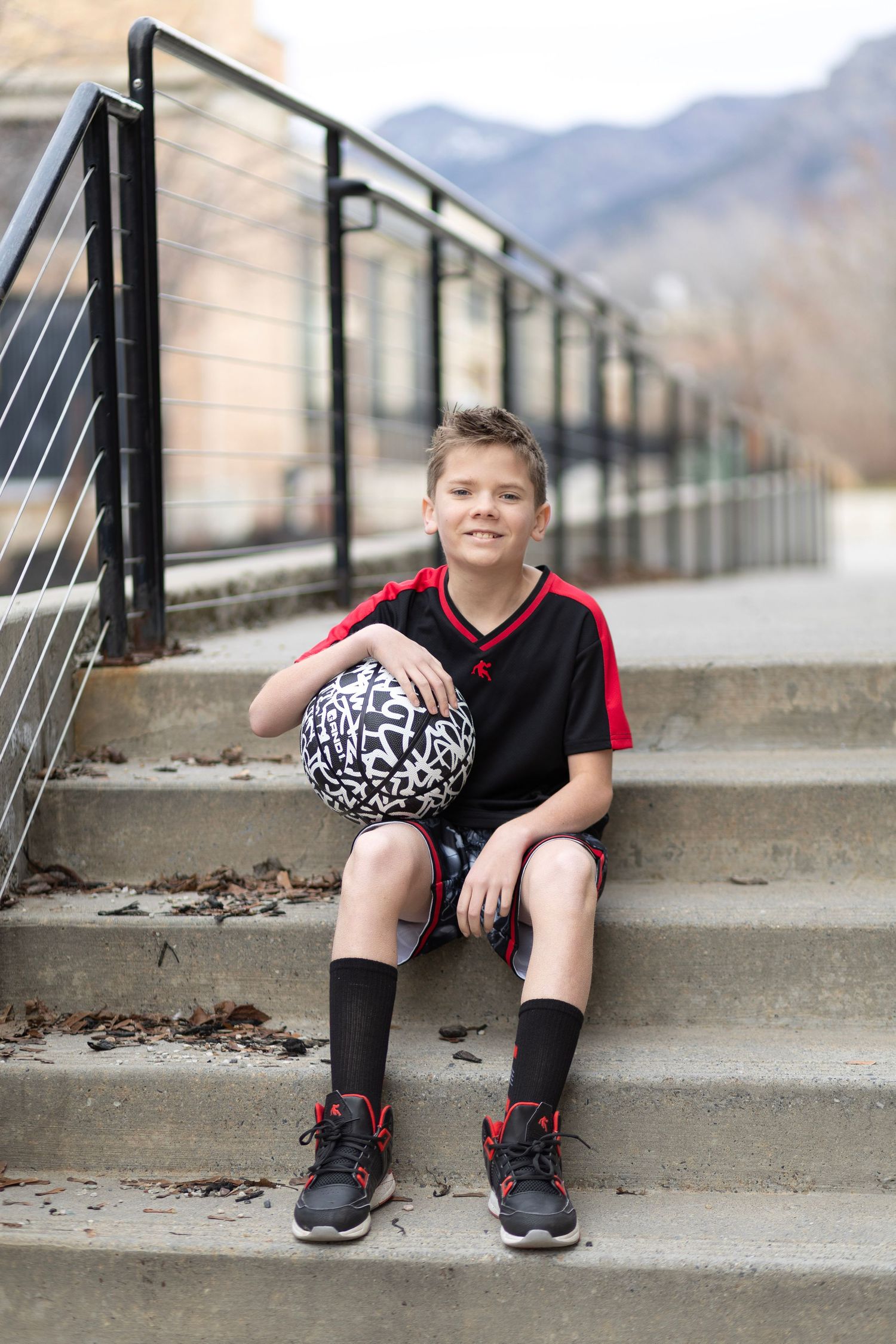 A young boy in a black soccer jersey sits on outdoor steps holding a patterned ball, wearing black socks and sneakers.