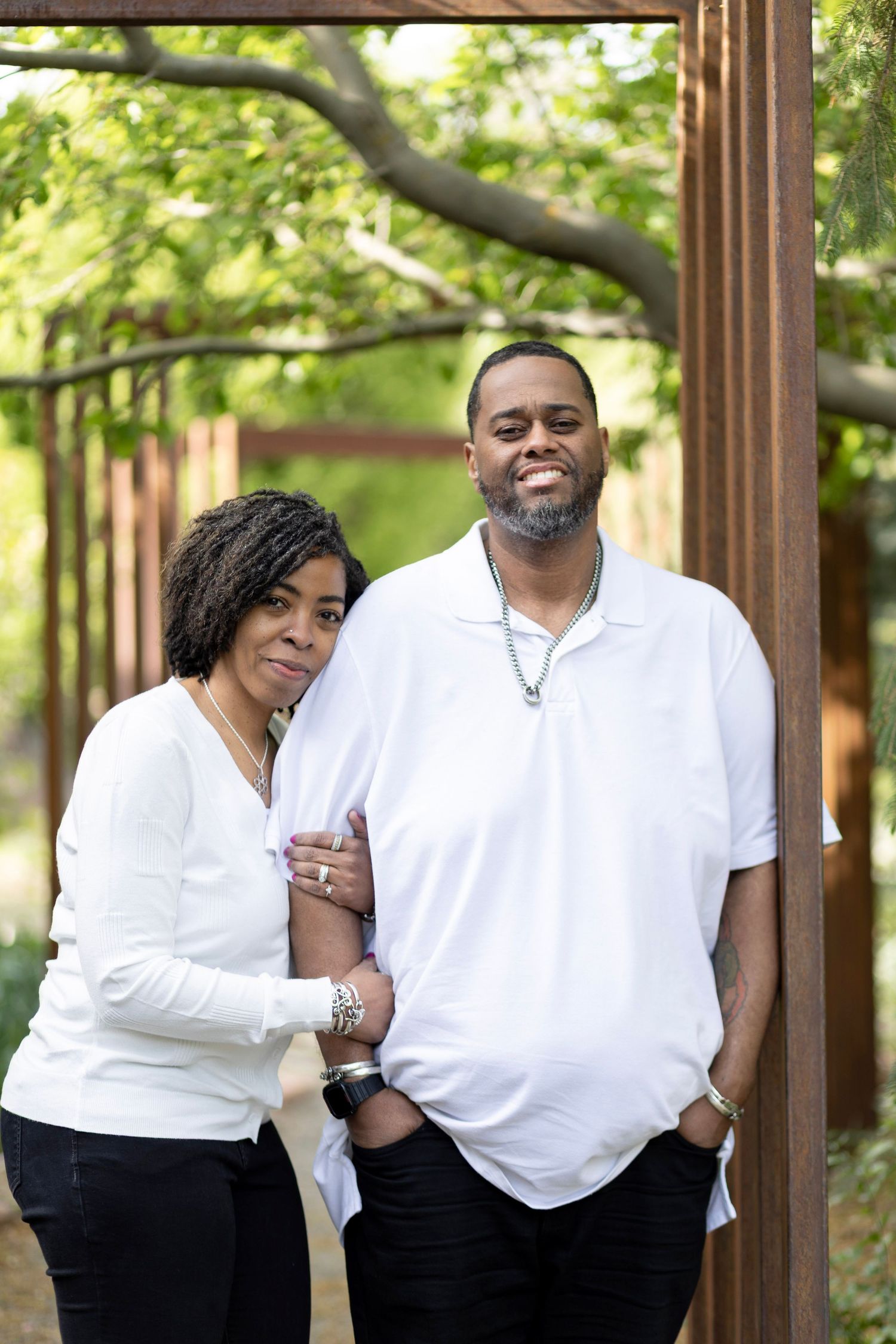 A smiling couple in matching white tops and black pants poses outdoors near a rustic metal pergola with green trees.