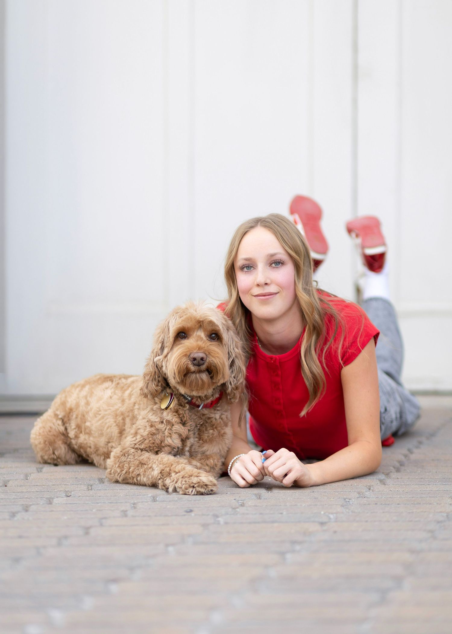 Young woman in red shirt lying on pavement with fluffy golden cockapoo dog, smiling outdoors.