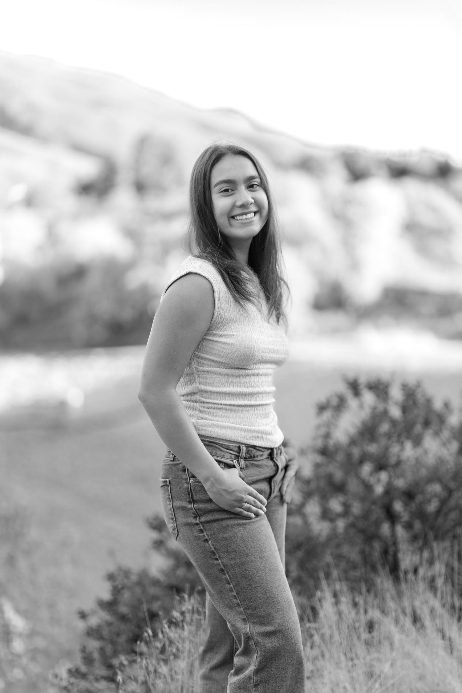 A person in a white tank top and jeans poses outdoors in a field with greenery and natural lighting.