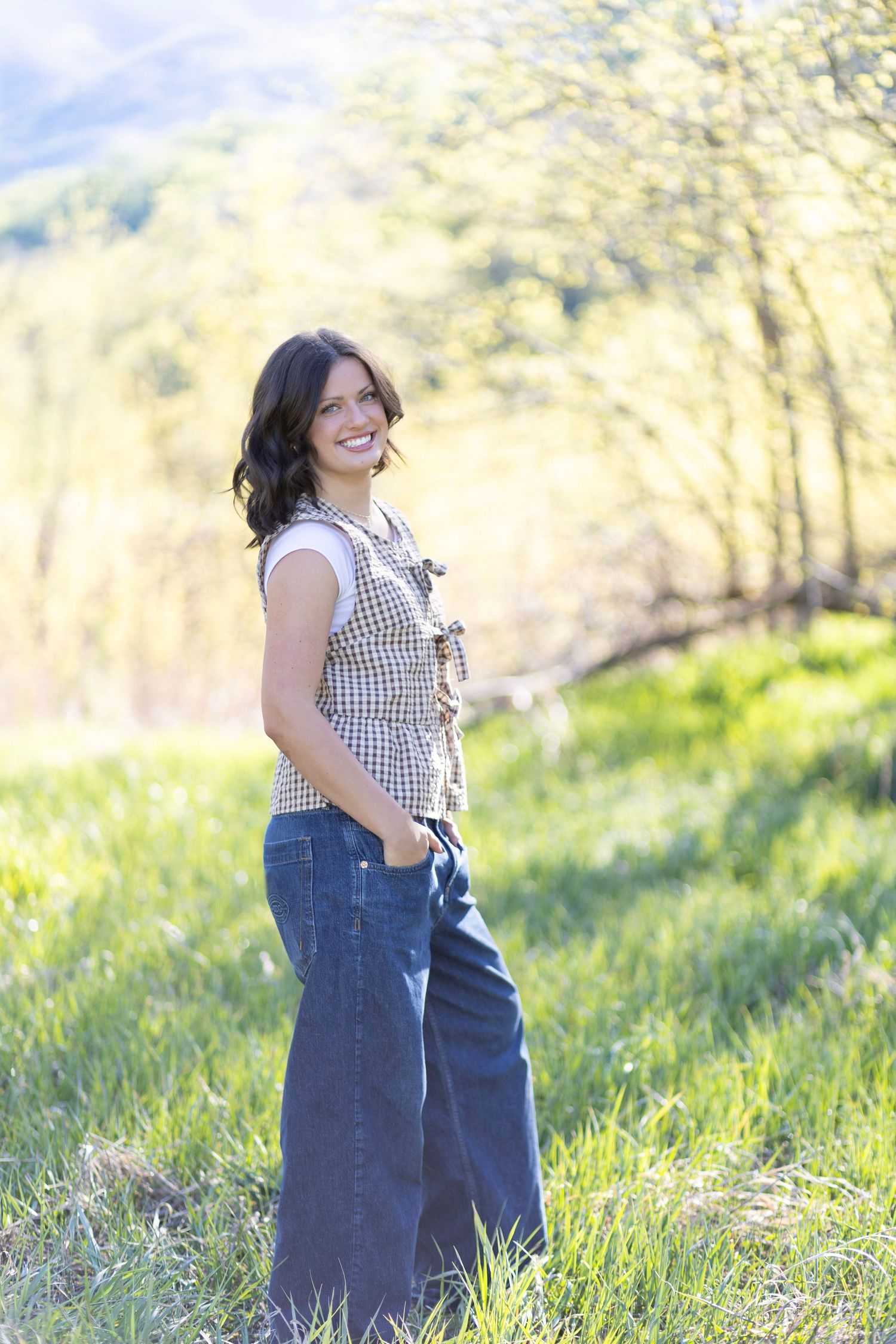 Smiling woman in plaid top and wide-leg jeans standing in a sunny green meadow with trees in background.