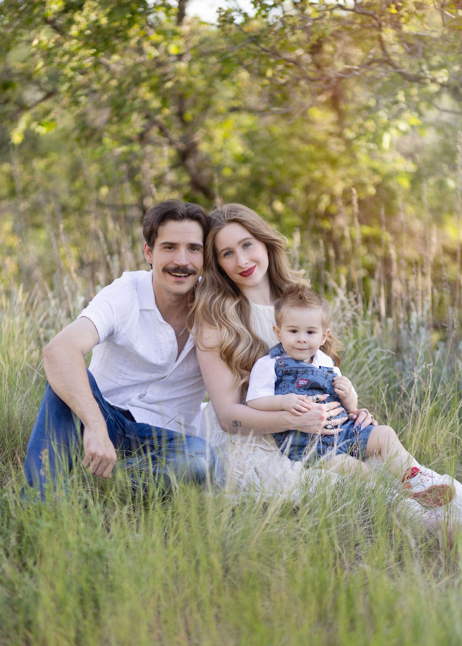 Family of three sitting in lush green meadow, parents smiling with toddler in denim overalls on sunny day.
