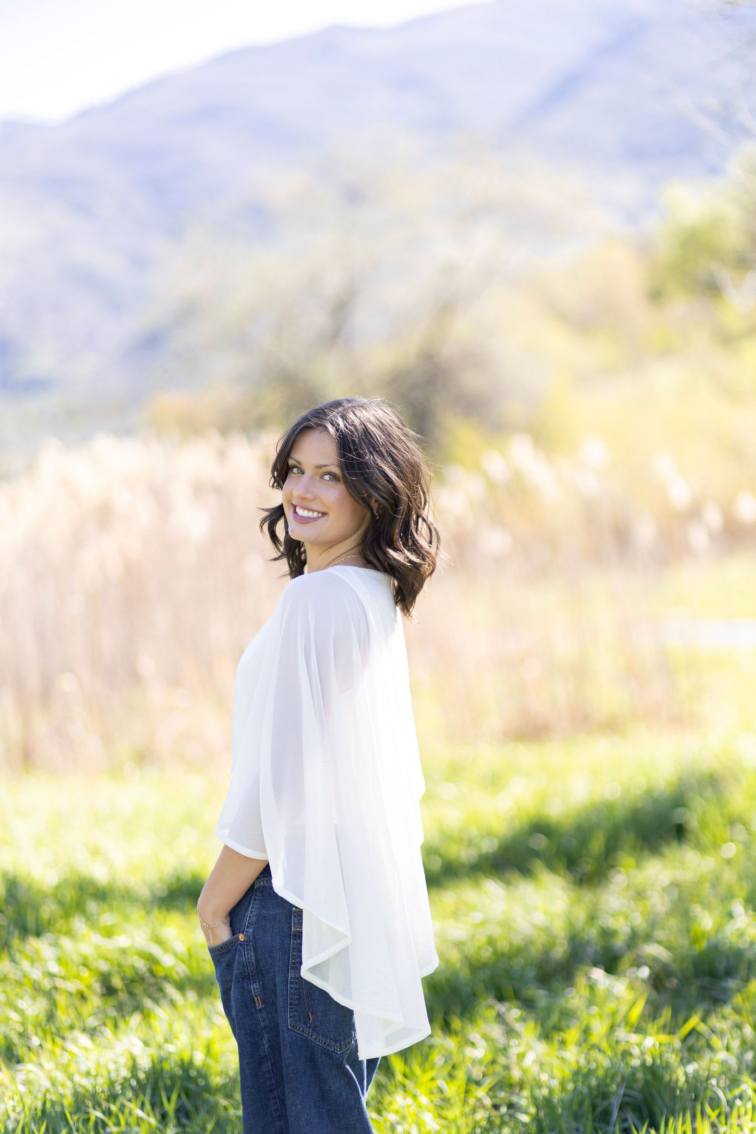 Smiling brunette woman in white blouse and jeans standing in a sunny green field with mountains in the background.