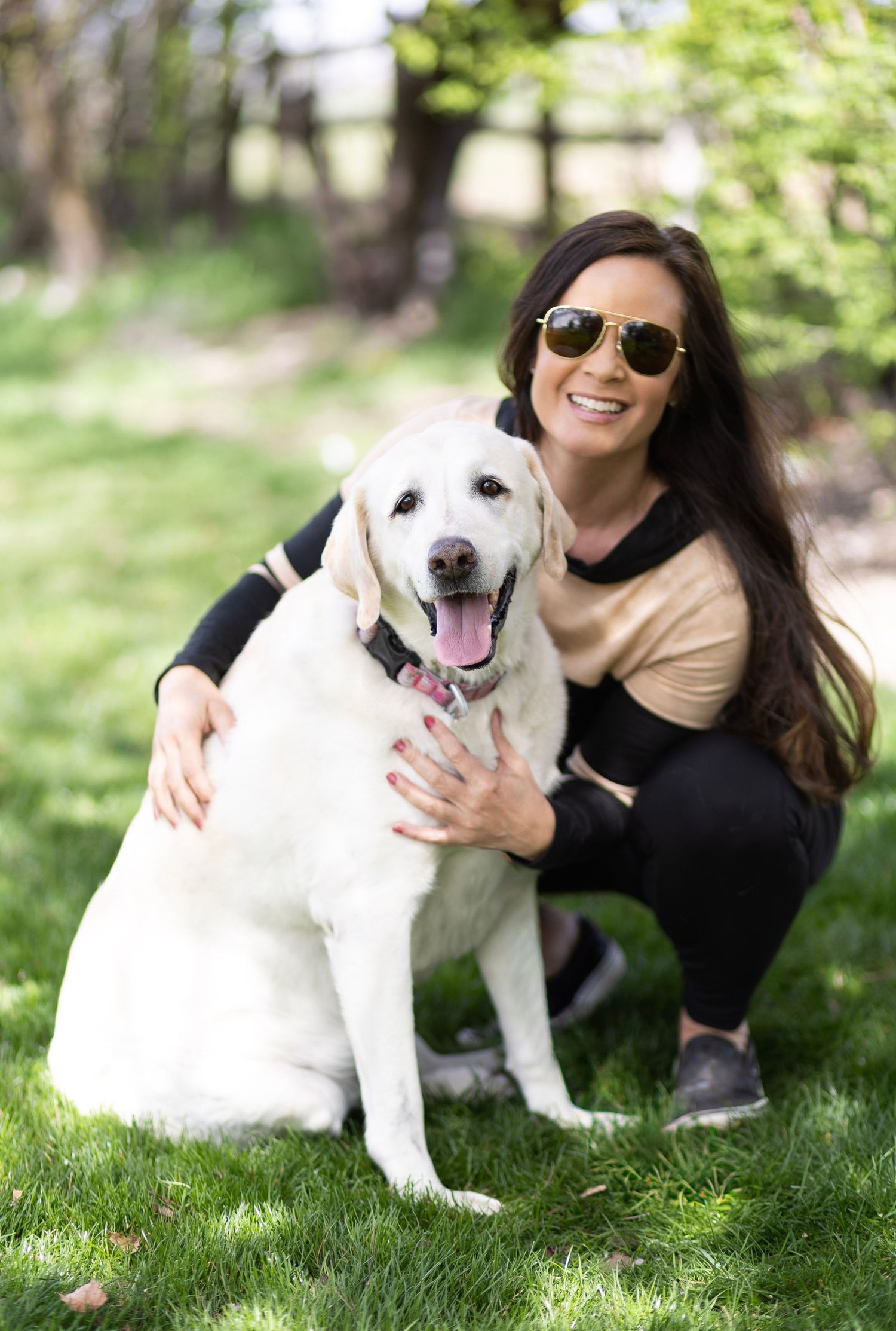 Brunette woman in sunglasses crouching beside a white Labrador Retriever dog on a sunny lawn.