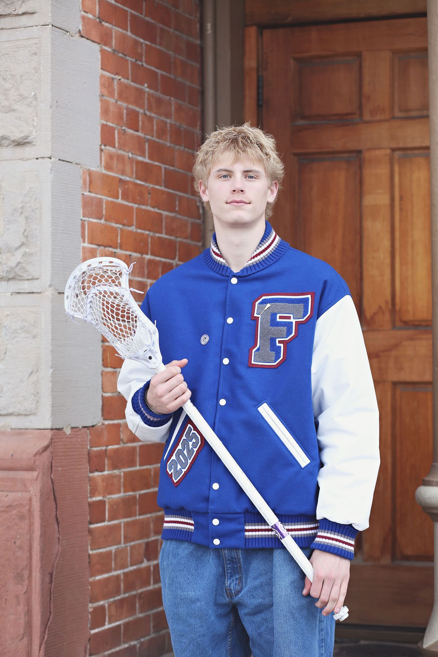 Someone holds lacrosse sticks while wearing a blue varsity jacket against a brick wall.