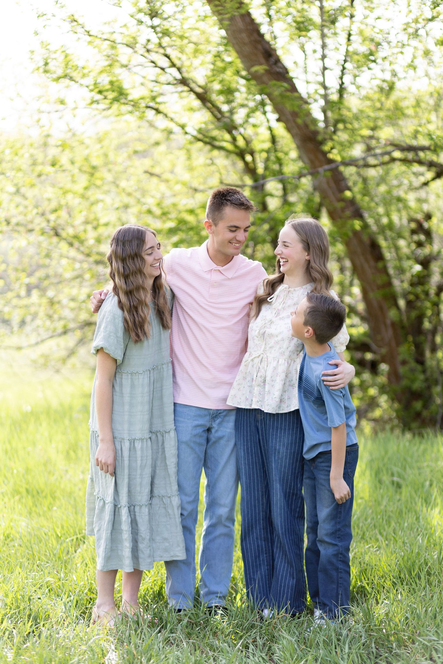 Four happy siblings laughing together outdoors in a green park setting on a sunny day.