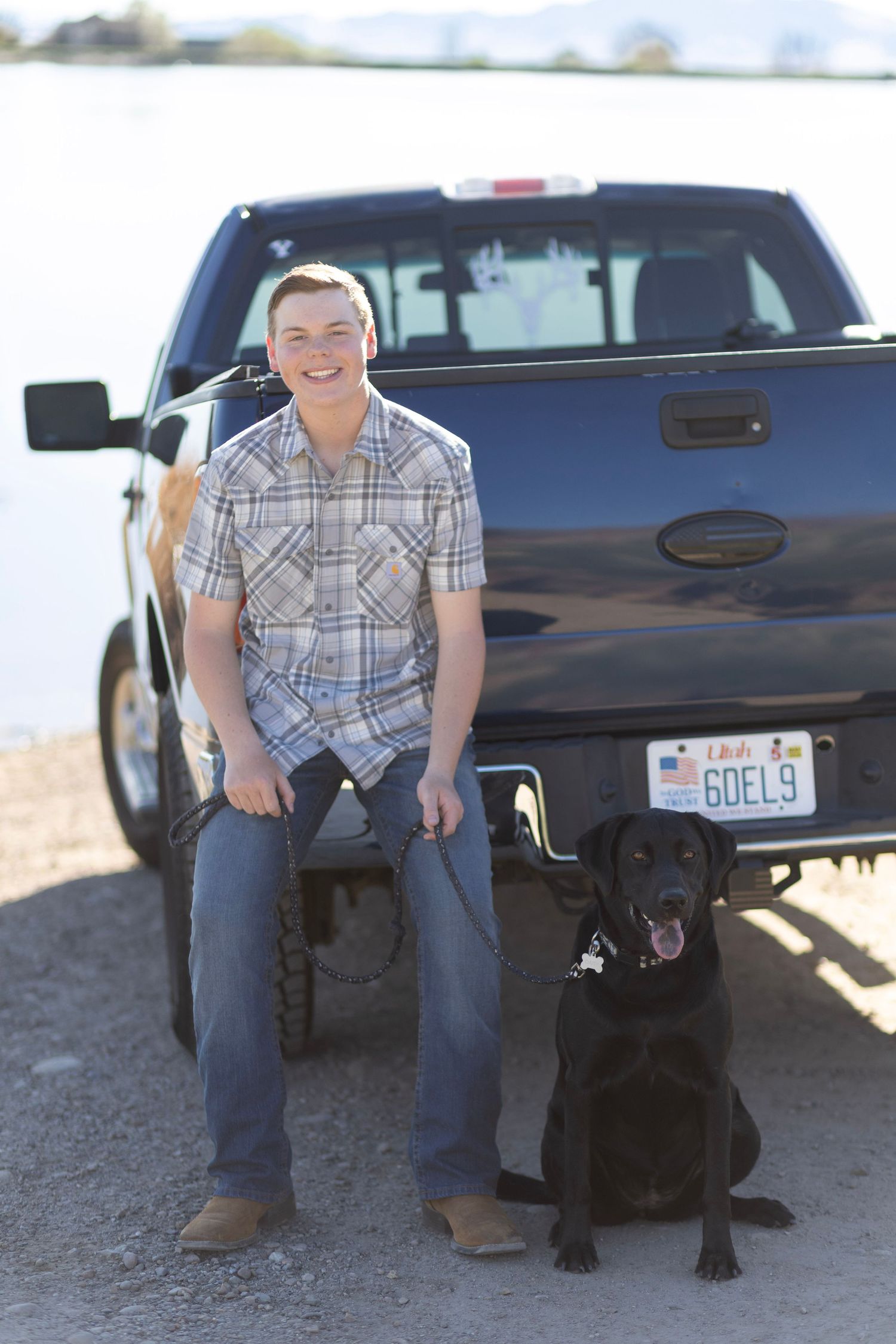 Young man smiling beside black pickup truck with black Labrador dog near lakeside on sunny day.