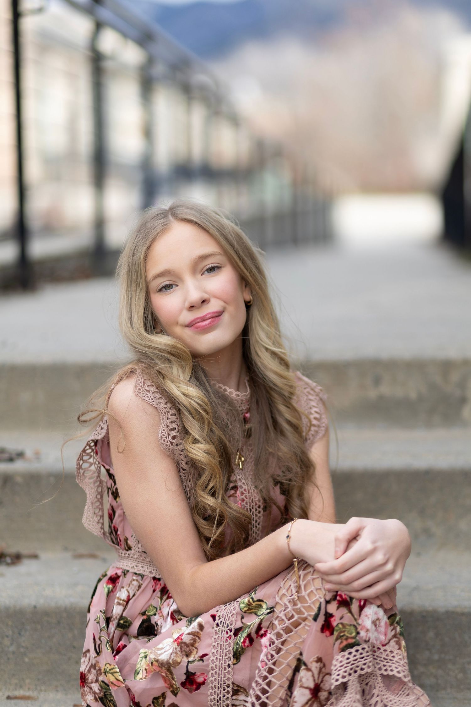 Young girl with long blonde hair sits on steps wearing a pink floral dress, smiling softly outdoors.