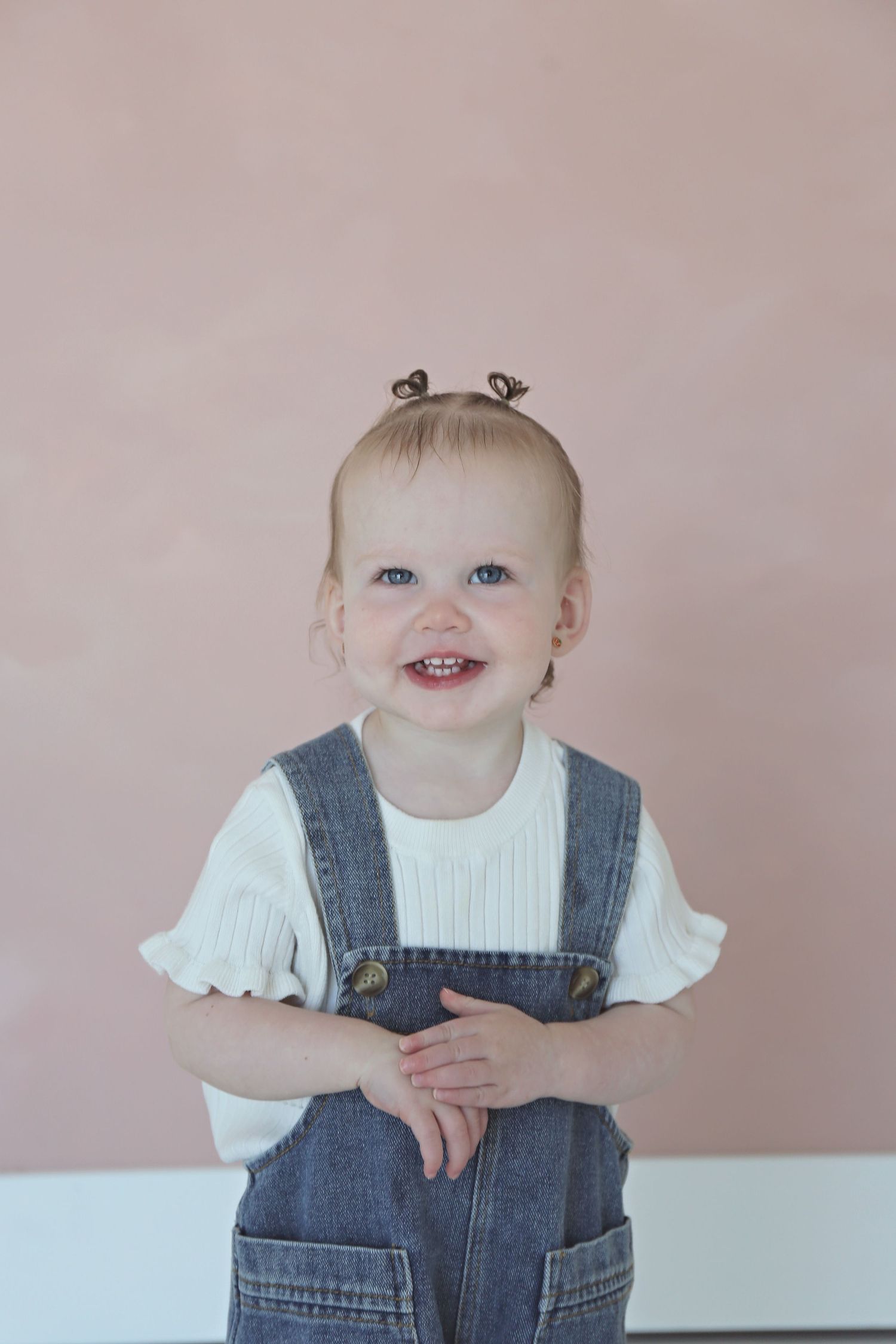 Smiling toddler in denim overalls and white shirt poses for studio portrait.
