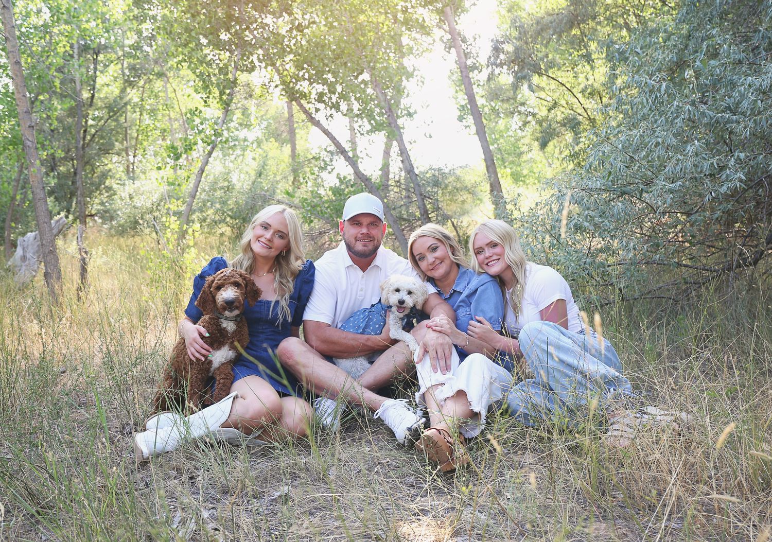 Family and pets relaxing together in a sunlit outdoor setting with trees in the background.