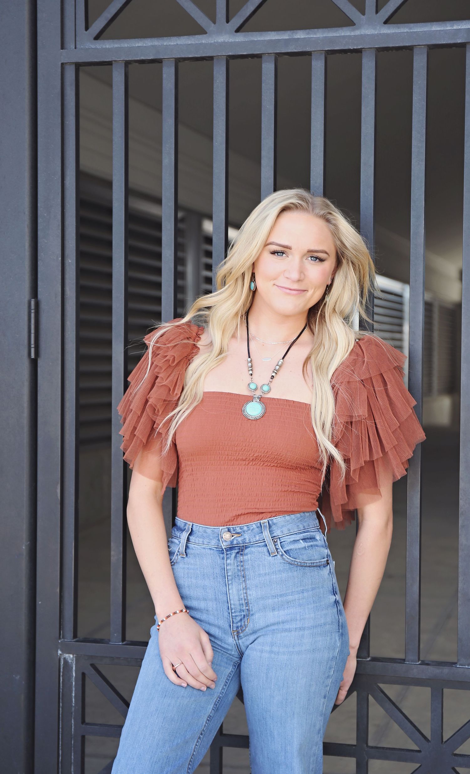 Person in rust-colored top and blue jeans posing against metal railings.