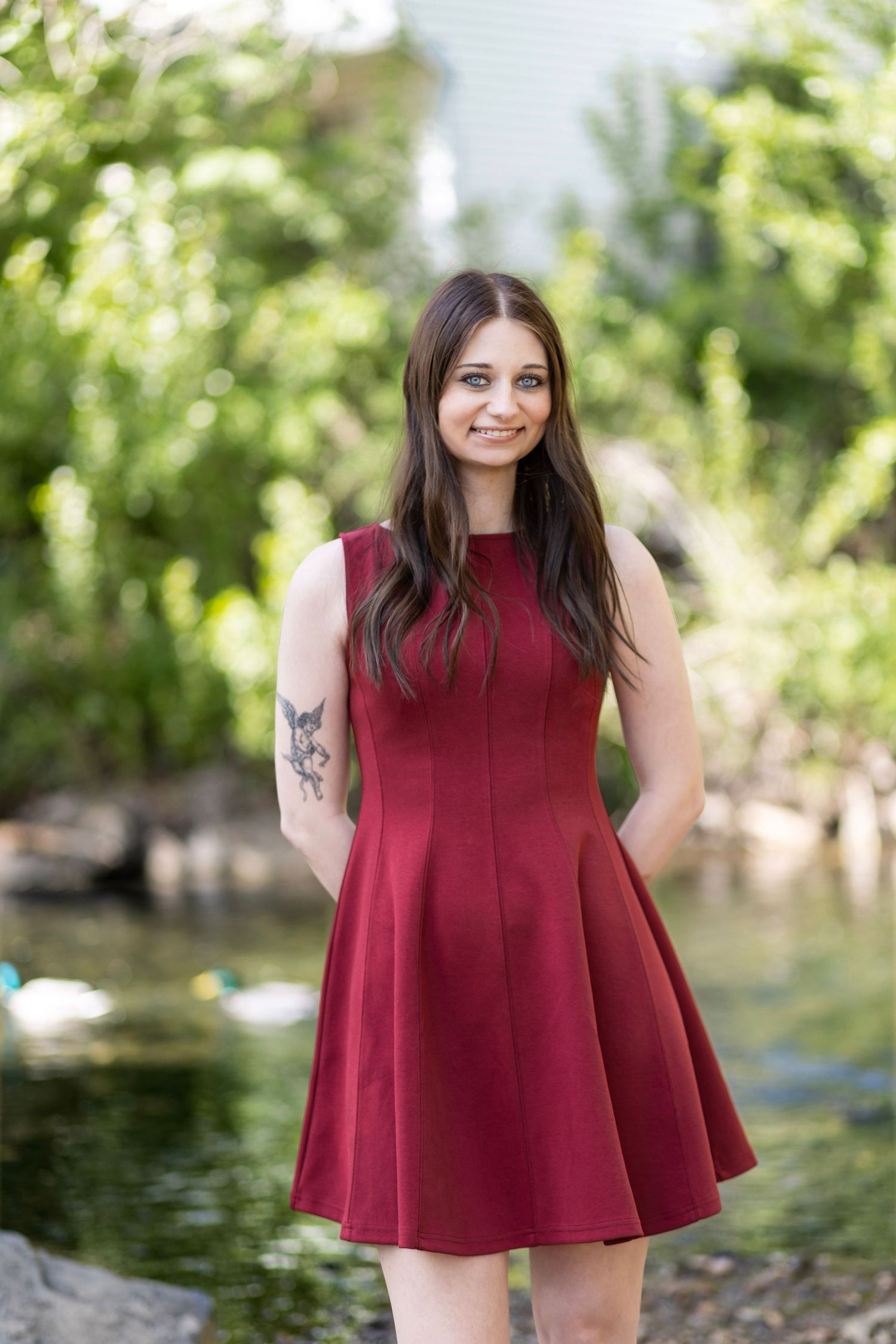 Young woman in a red sleeveless dress smiling outdoors near a pond with lush green trees in the background.