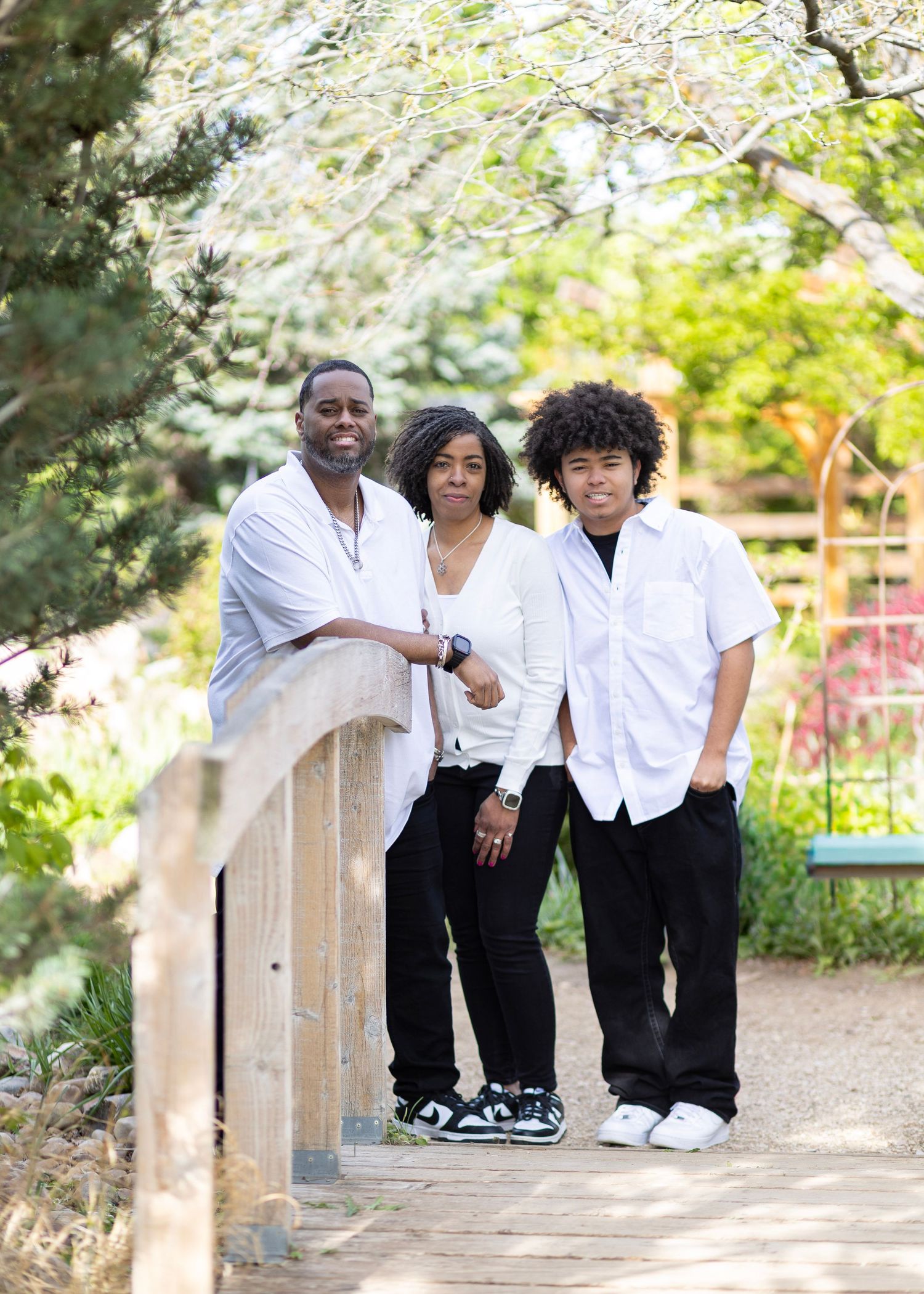 Family of three posing on a garden bridge, wearing matching white and black outfits, surrounded by lush greenery.
