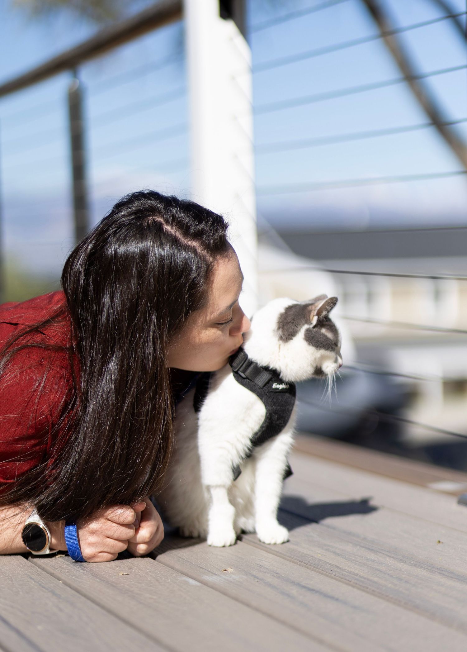 Woman in red top lying on deck, kissing her black and white cat wearing a harness outdoors.