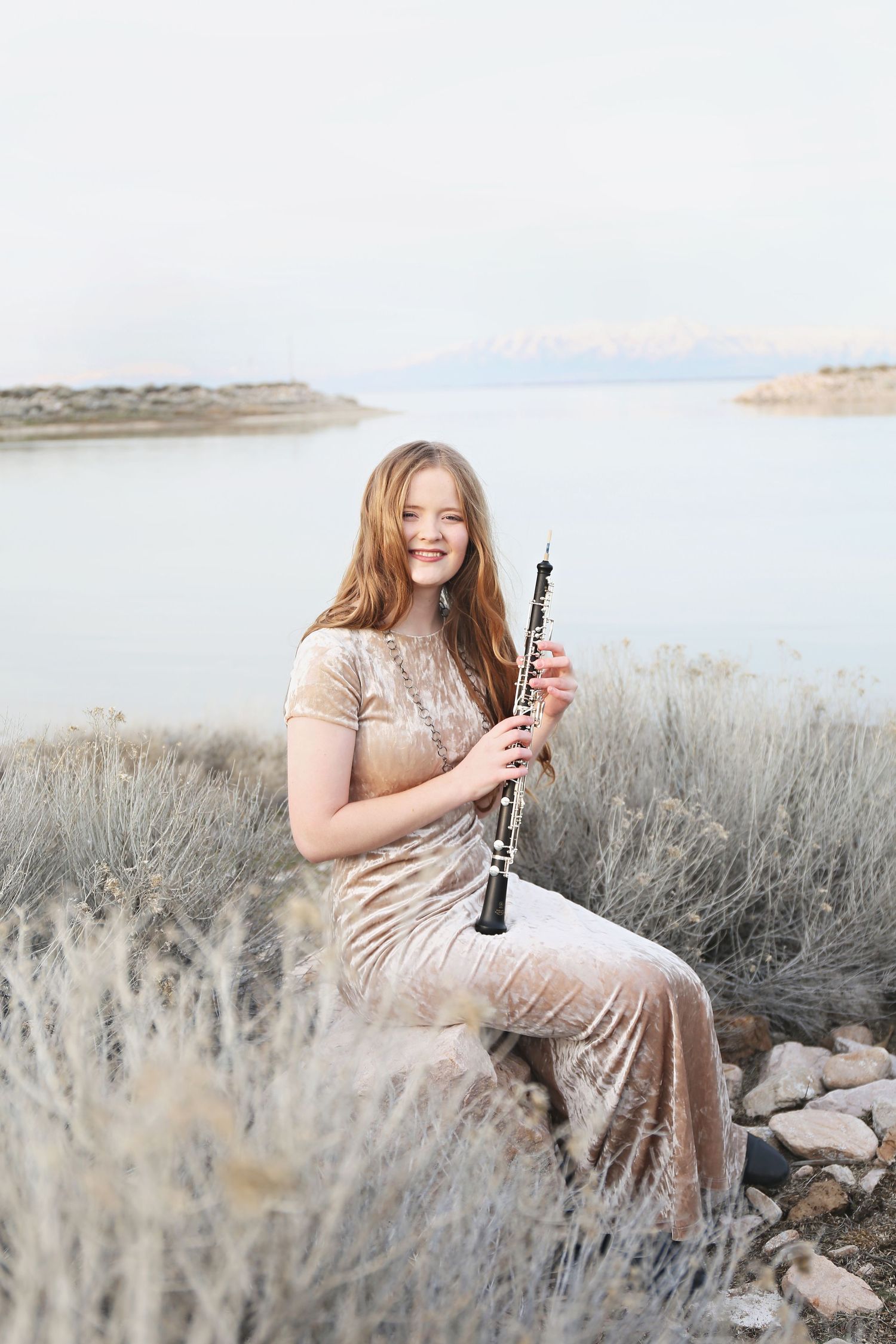 Person in sparkly dress holds clarinet while sitting among desert brush near lake.