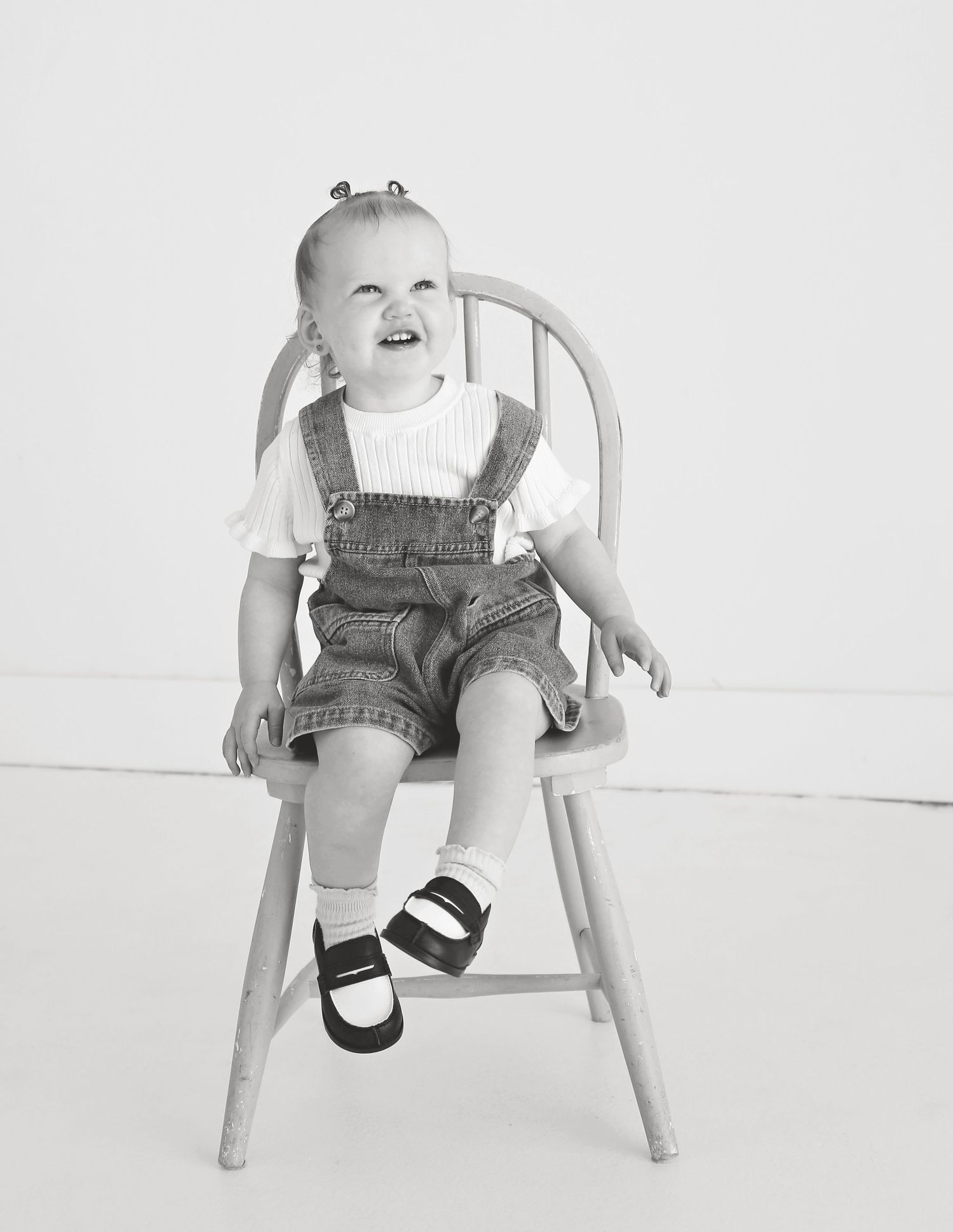 Black and white portrait of toddler in overalls sitting on vintage chair.