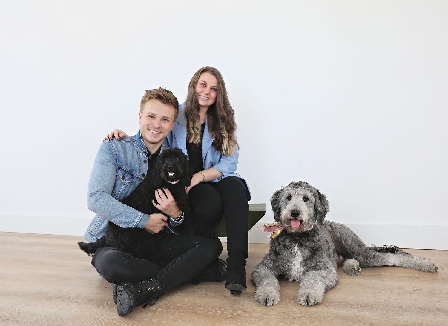 Two people sitting on wooden floor with a black puppy and gray shaggy dog in casual denim attire.