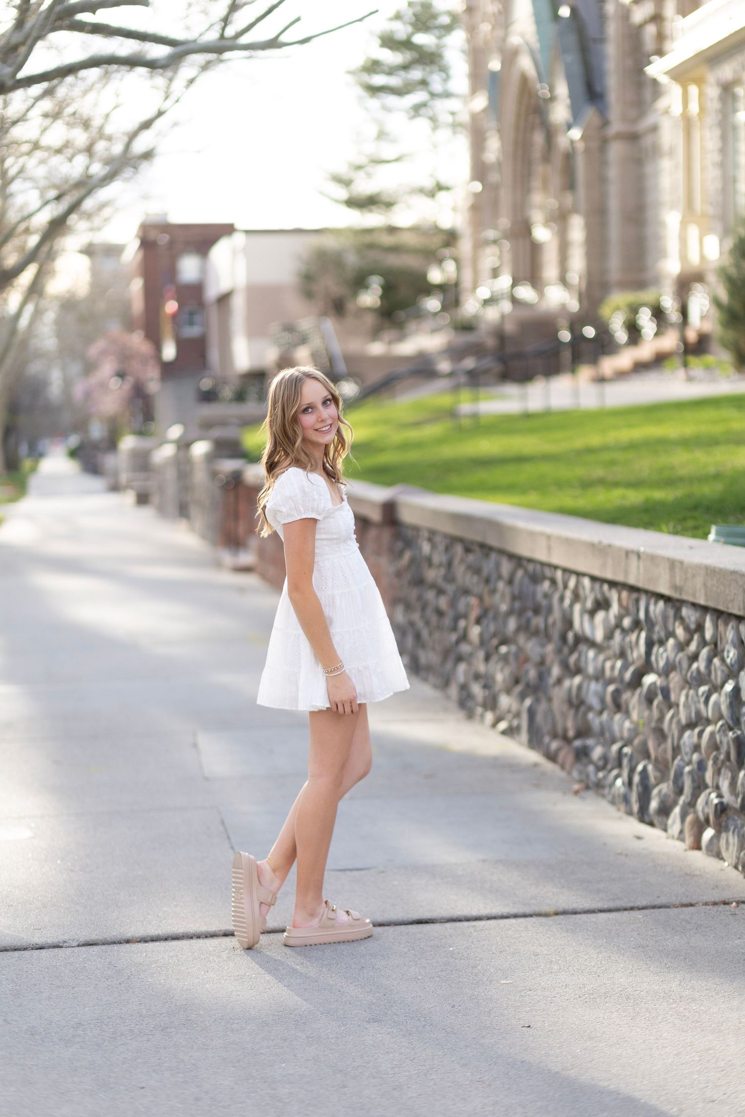 Young woman in white mini dress and pink sneakers posing on a sidewalk near a stone wall on a sunny day.