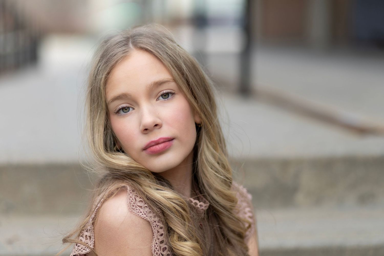 Young girl with long blonde hair wearing a pink lace dress, posing outdoors on a city street.