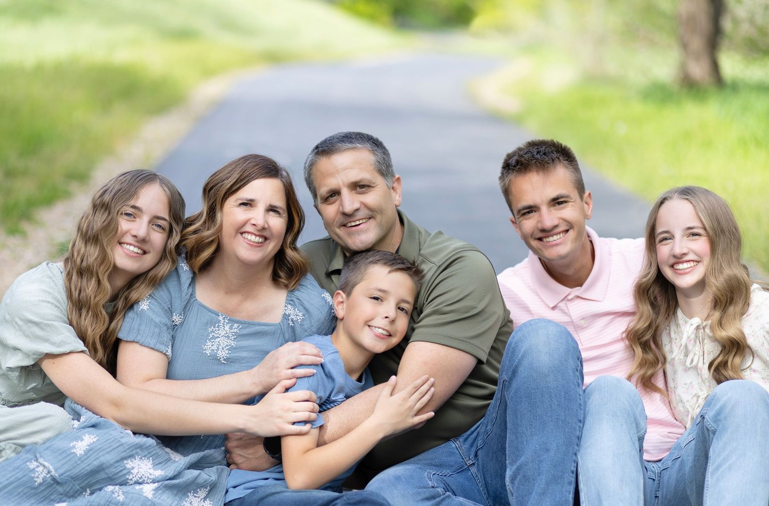 Happy family of six smiling together outdoors on a tree-lined path on a sunny day.