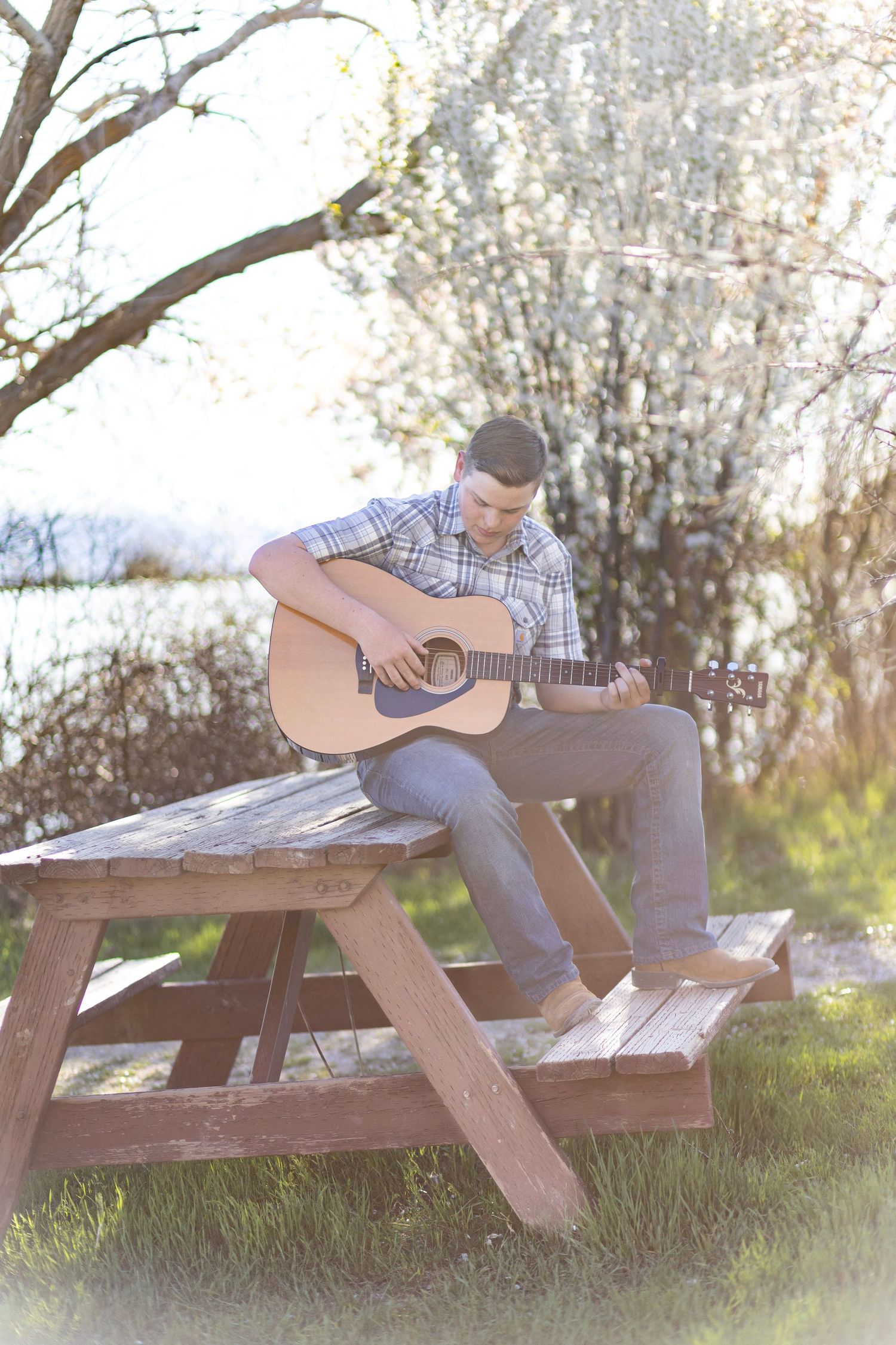 Young man playing acoustic guitar while sitting on a wooden picnic table outdoors near a serene lake.