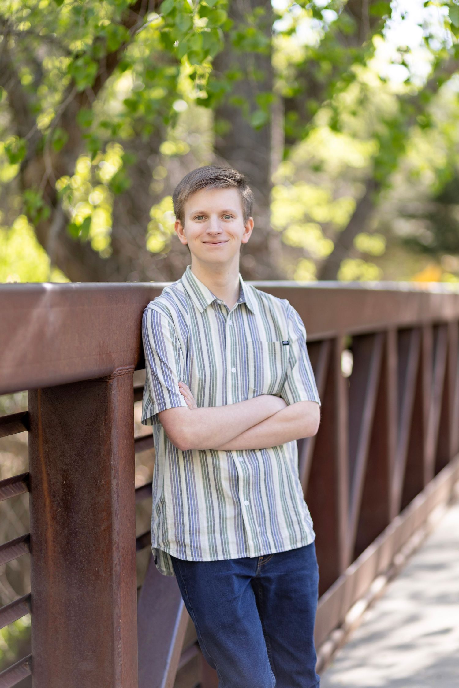 Young man in striped shirt leaning on rustic bridge railing, smiling with arms crossed, surrounded by green trees.