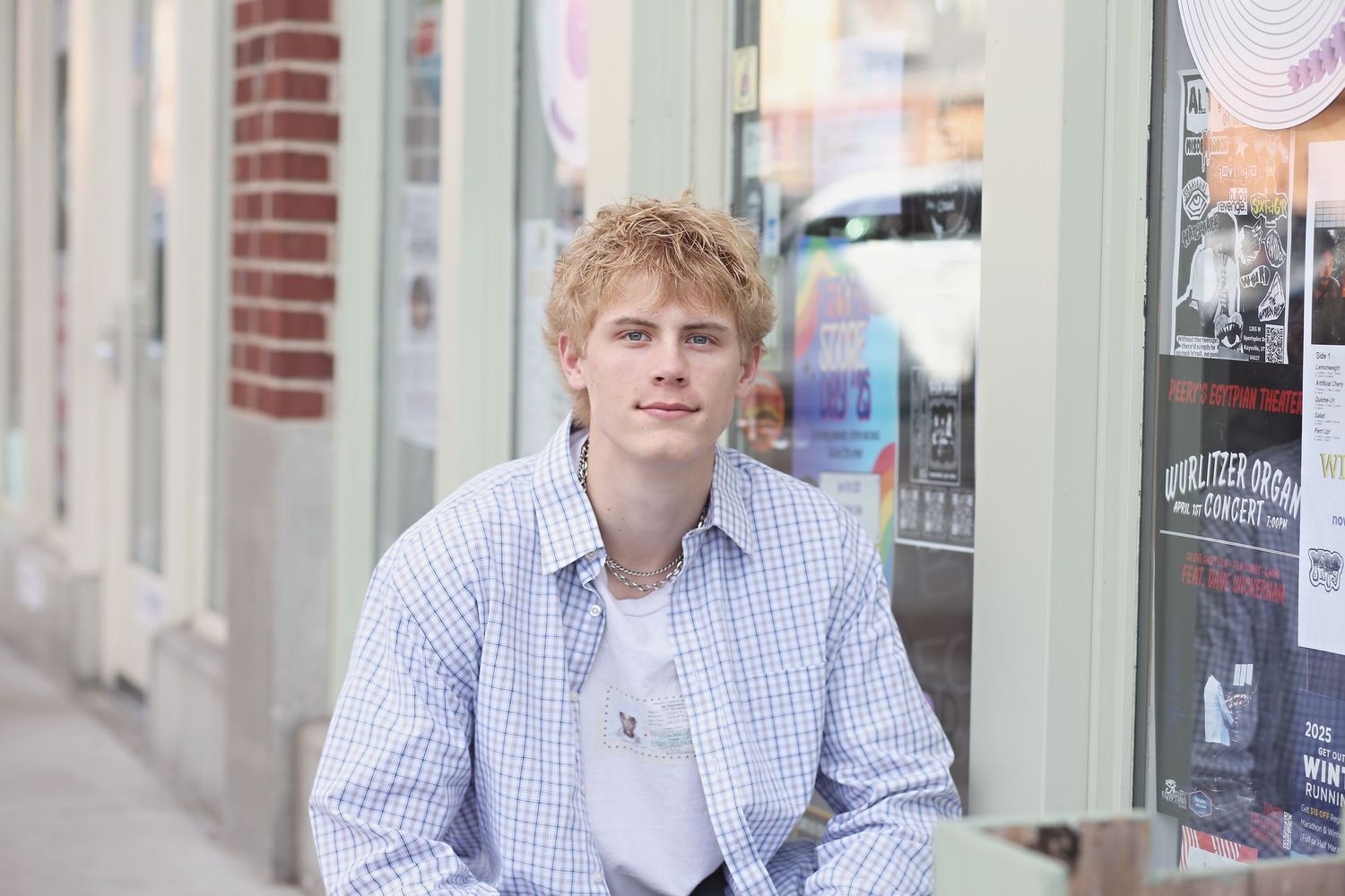A person in a light blue striped button-down shirt and blonde hair stands against a brick wall background.