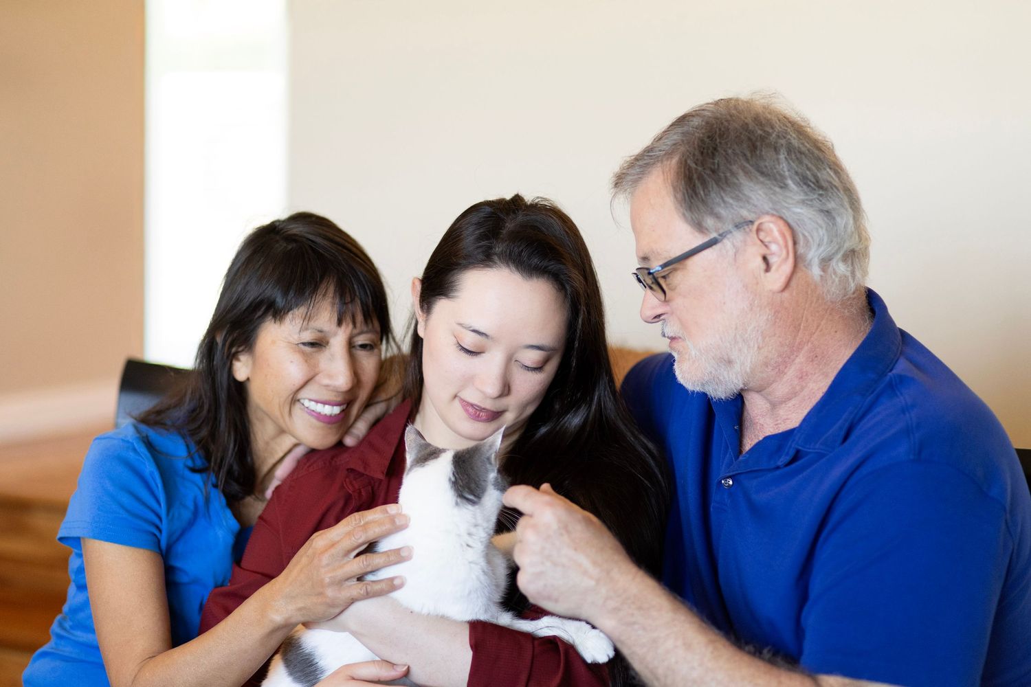 Family of three smiling adults petting a gray and white cat indoors.