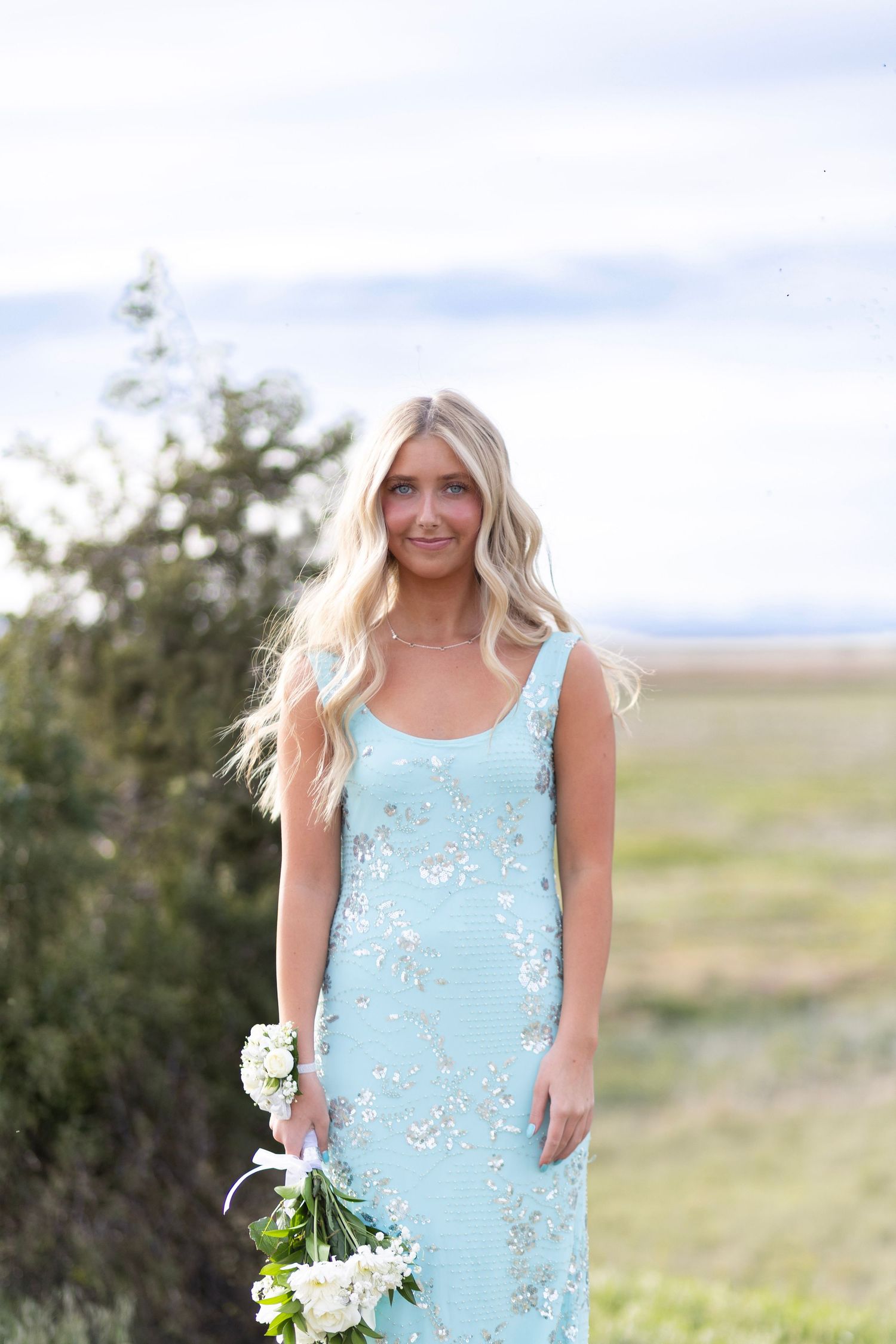 Blonde woman in a mint green sequined dress holding white flowers outdoors in a scenic field.