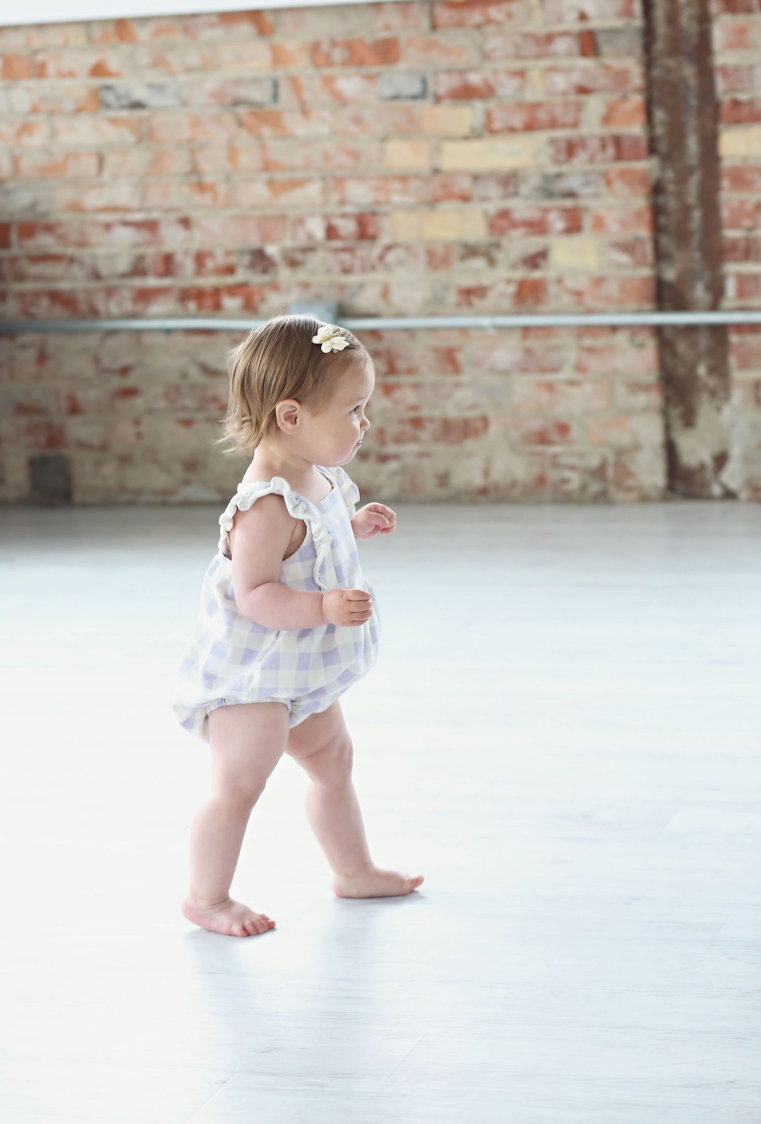 A toddler in a ruffled outfit takes steps across a bright indoor space with brick walls.