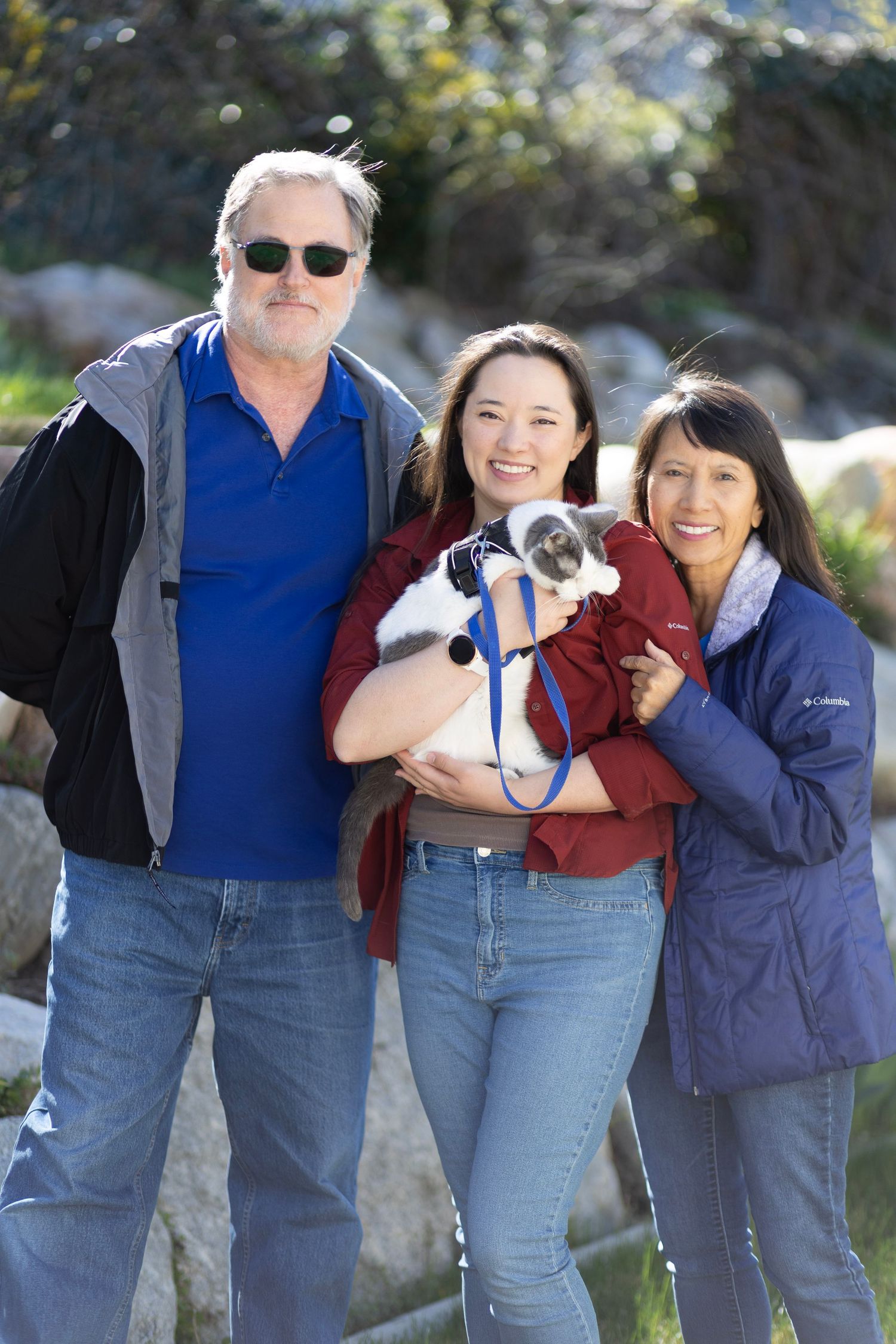 Two women and a man posing outdoors holding a small black and white cat on leash.