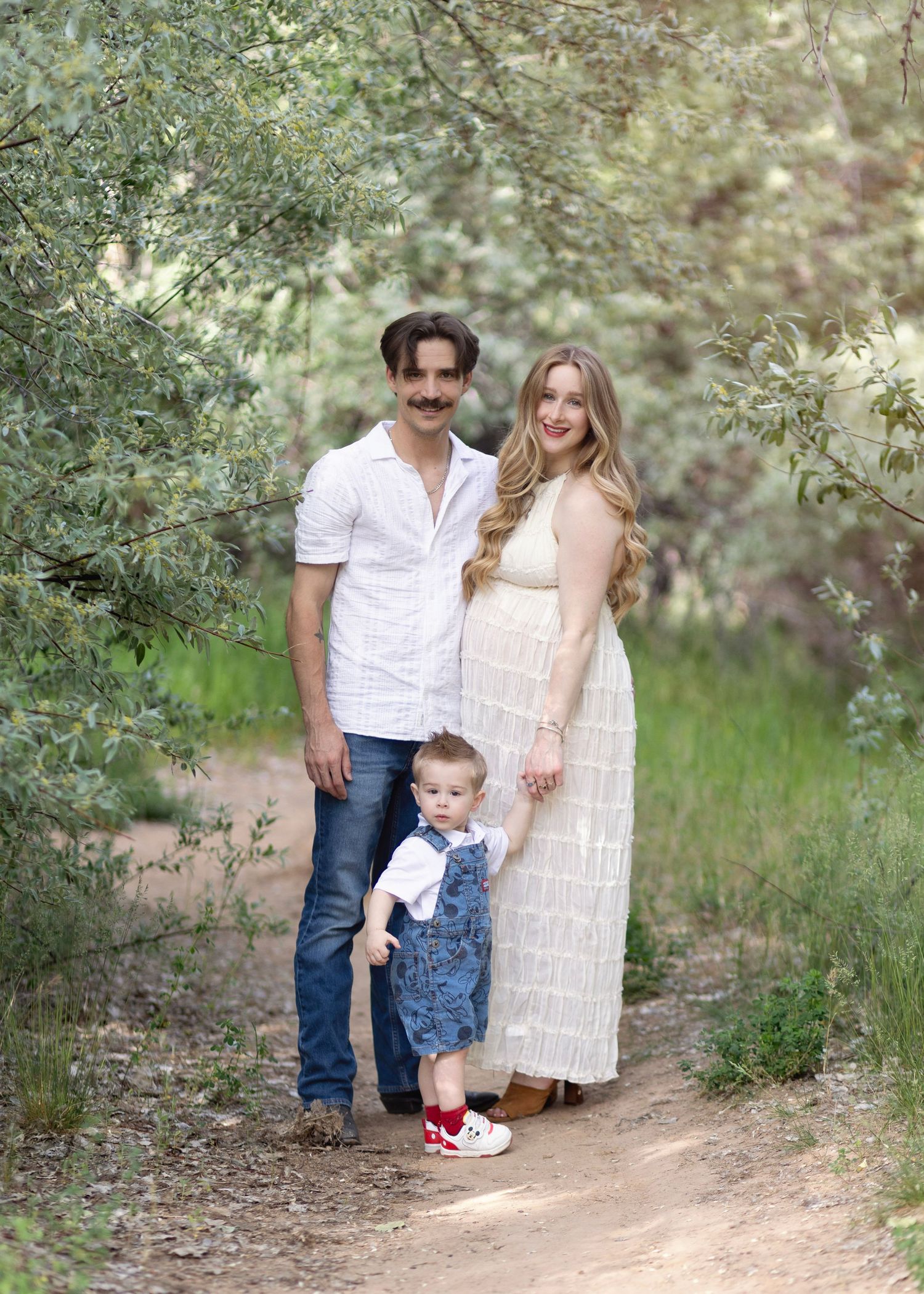 Pregnant woman and man pose with toddler on a nature path surrounded by lush green trees.
