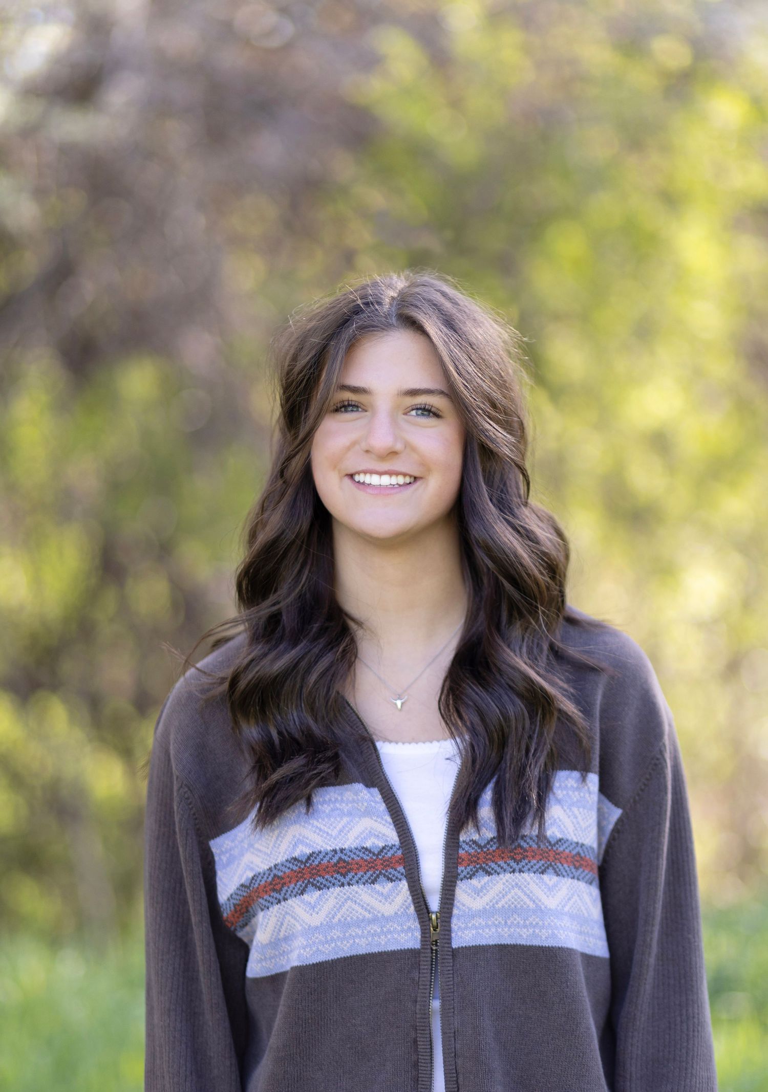 Young woman with long dark hair smiling outdoors in a gray patterned zip-up jacket among green trees.