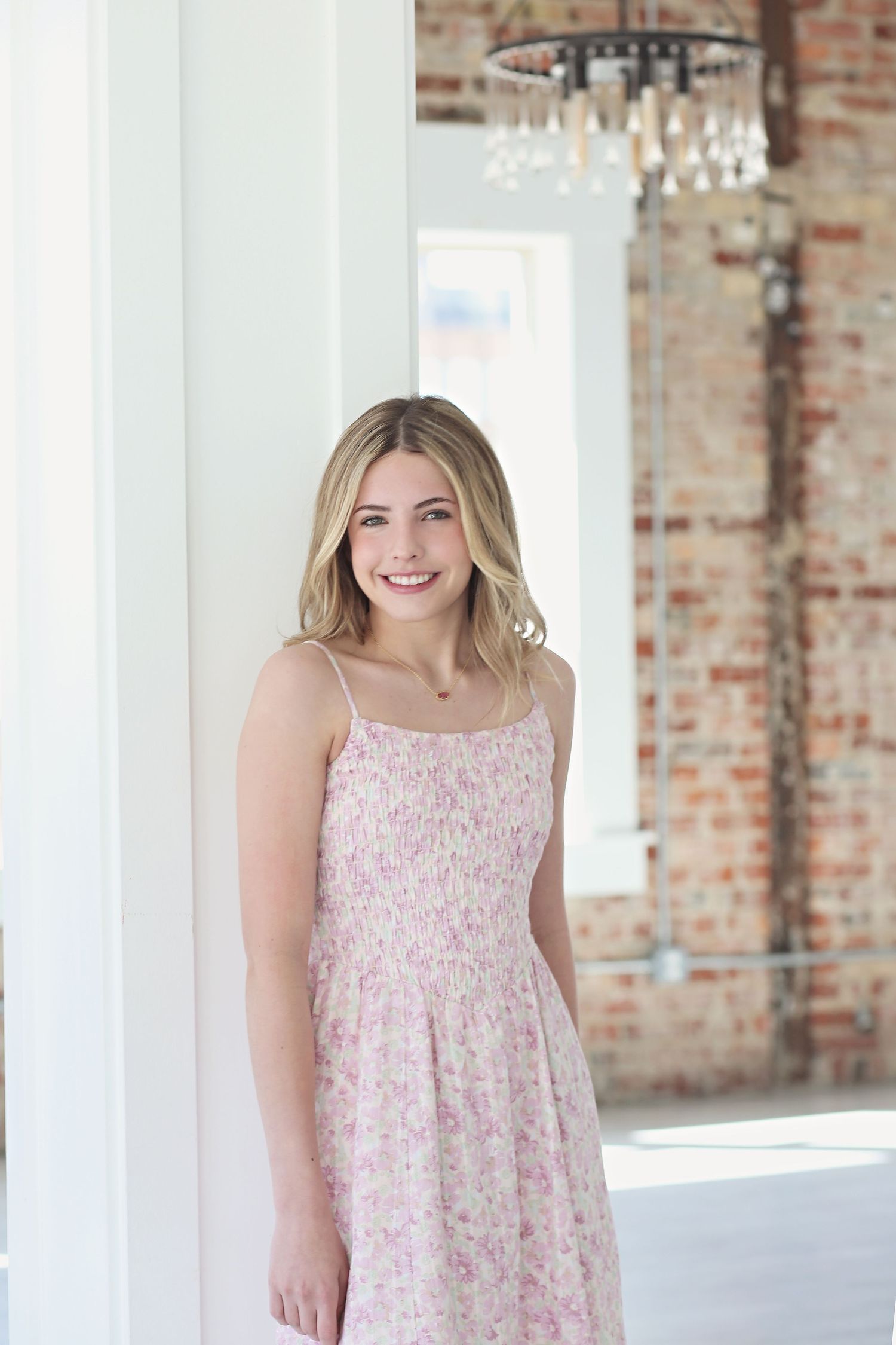 Young woman in floral pink dress standing in front of exposed brick wall with white columns.