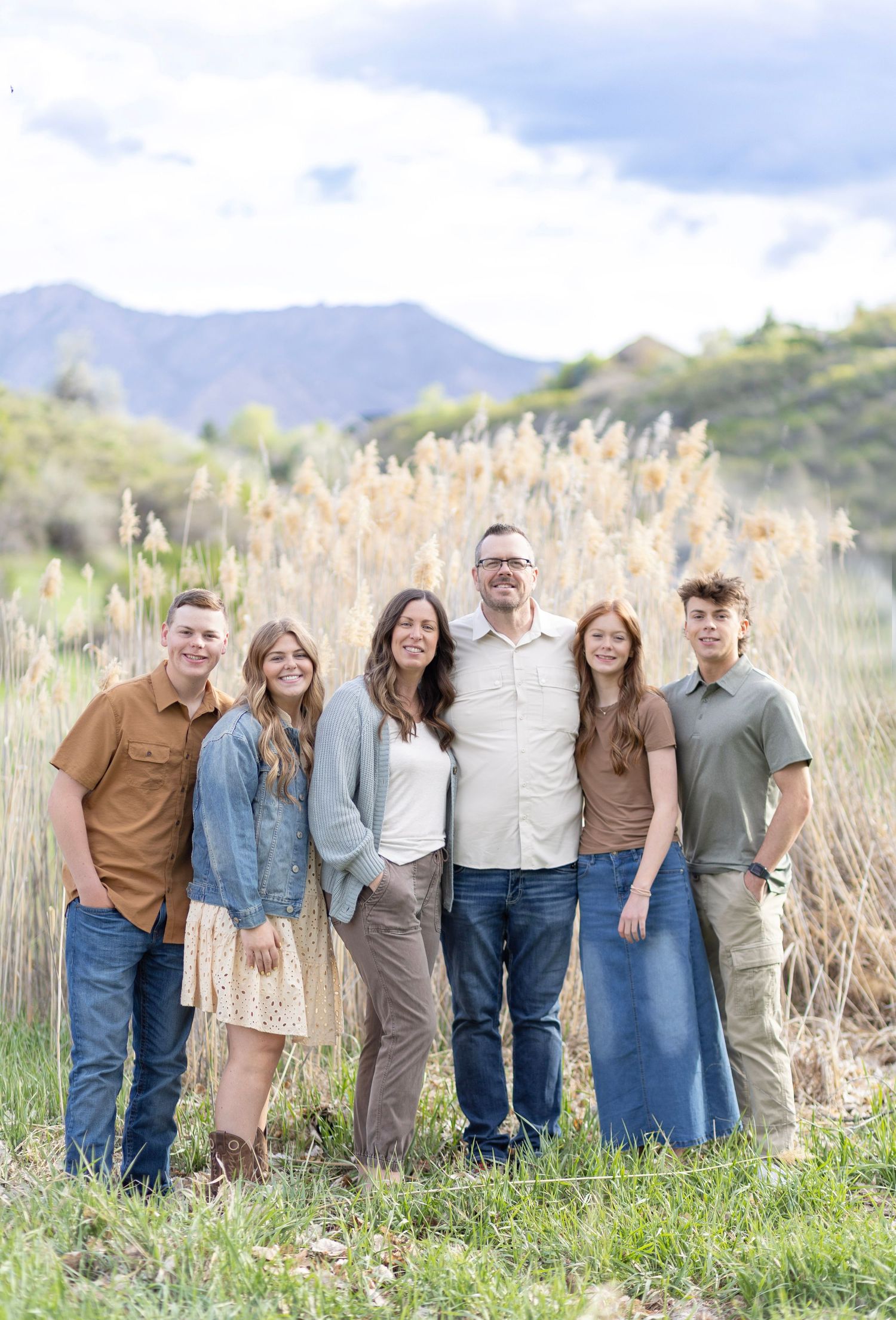 Family of six smiling outdoors with mountain backdrop and tall golden grasses.