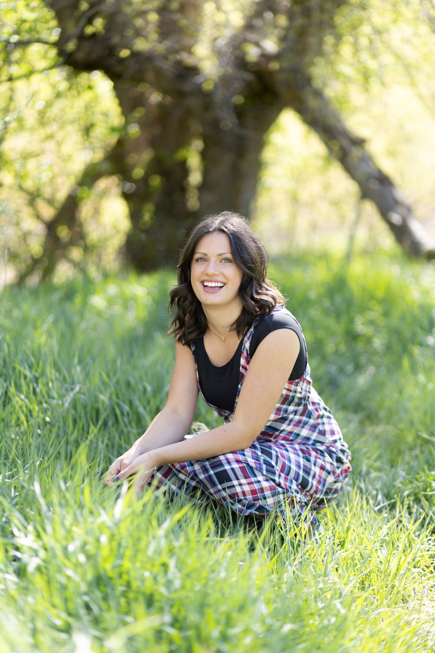 A smiling brunette woman sits in lush green grass wearing a plaid skirt, with trees in the background.