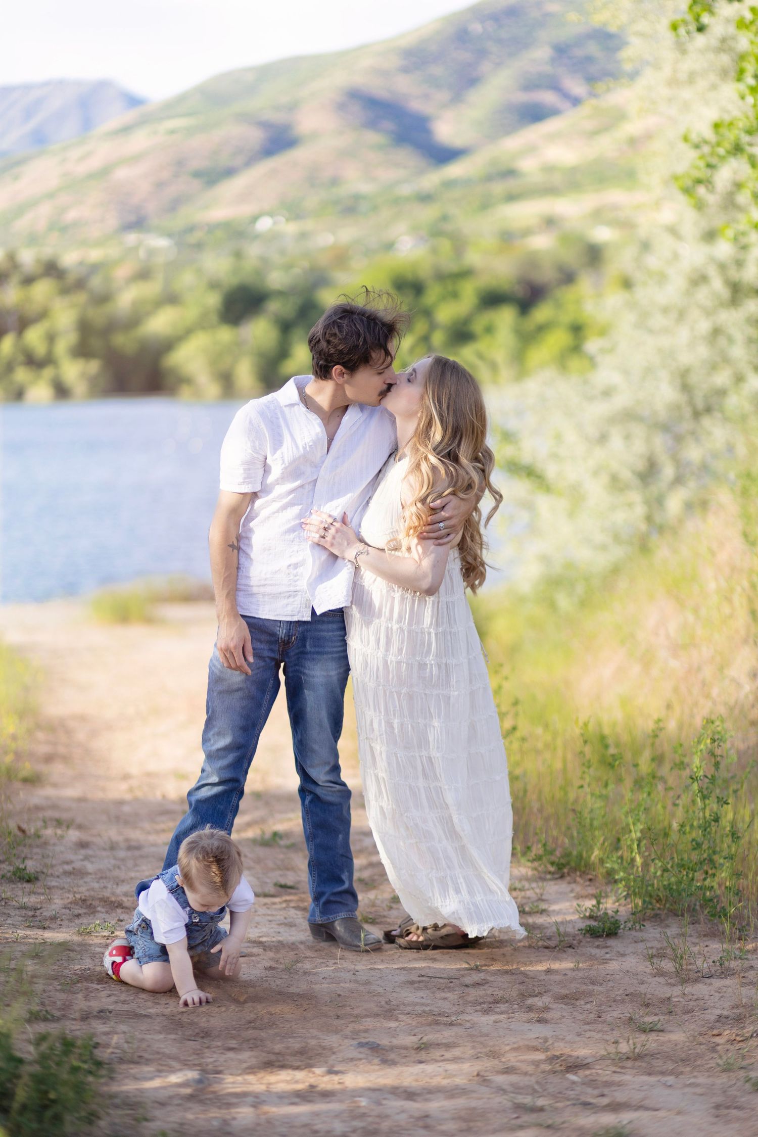 A couple kisses outdoors near a lake with mountains behind them while their toddler plays on a dirt path.