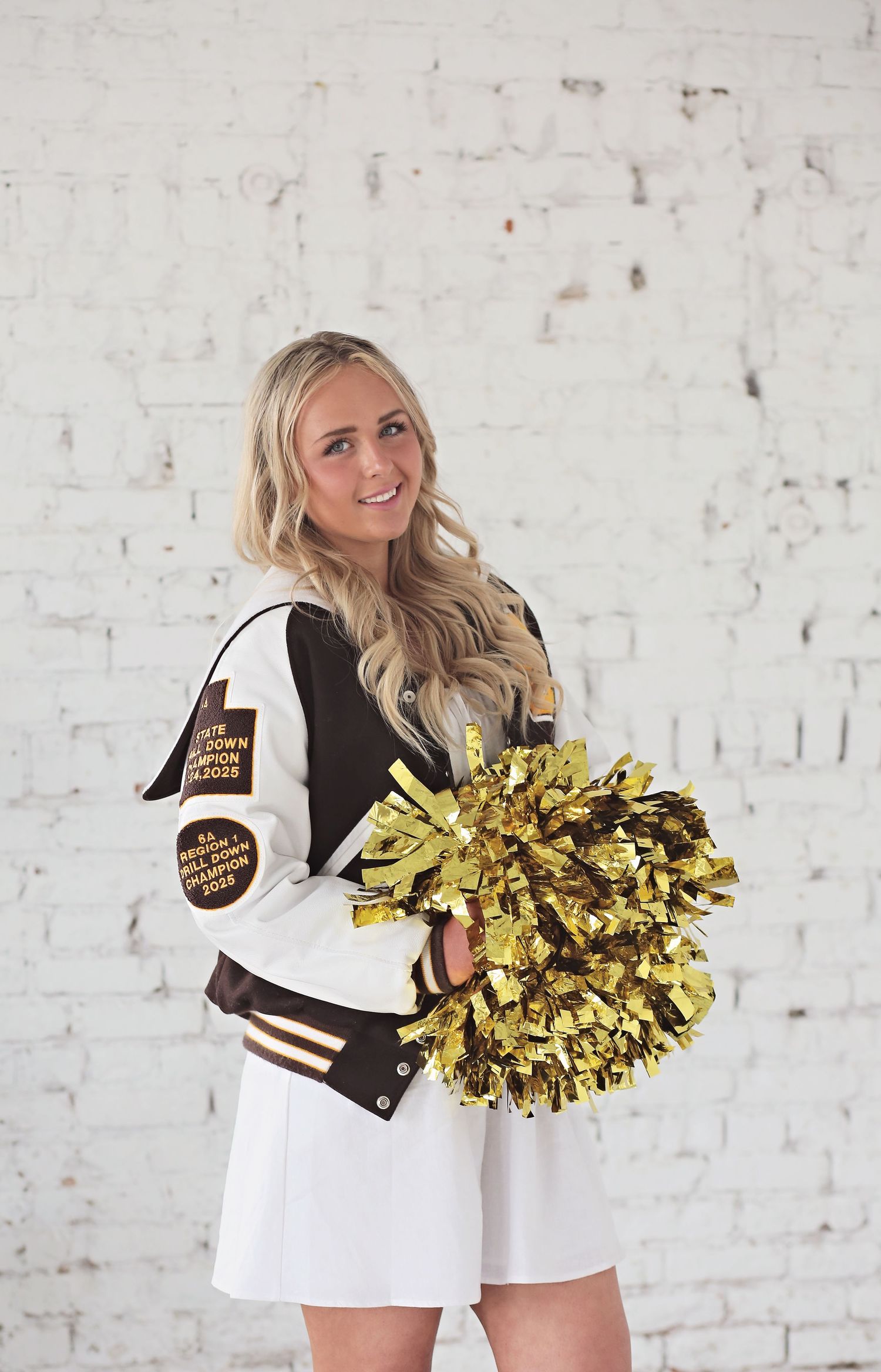 Cheerleader themed photoshoot with gold pom poms against white brick wall.